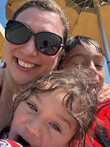 Smiling woman with sunglasses and two children posing for a close-up selfie under yellow beach umbrellas, capturing a joyful moment on their family travel bucket list.