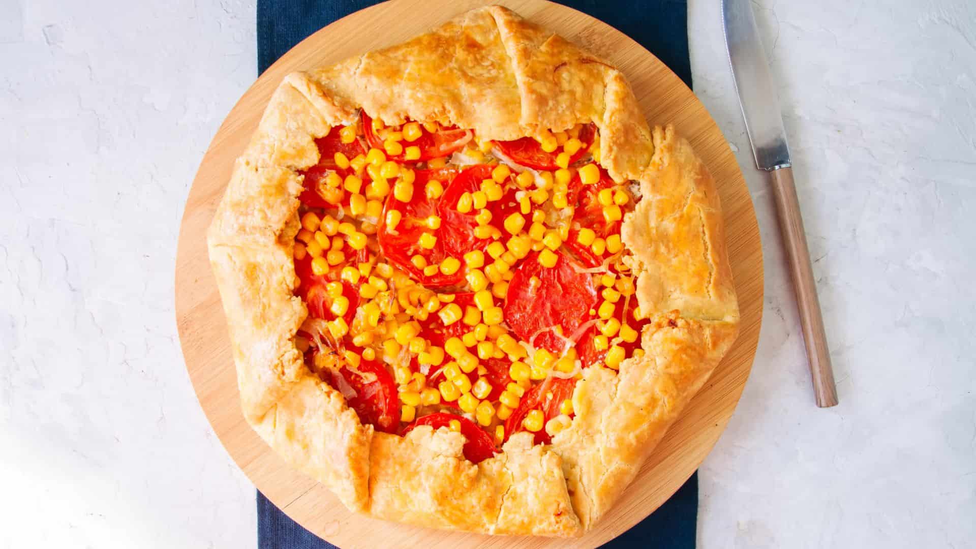 A rustic galette with tomatoes and corn on a wooden board, next to a knife&mdash;an inviting sample of the Best Vegetarian Food in France.