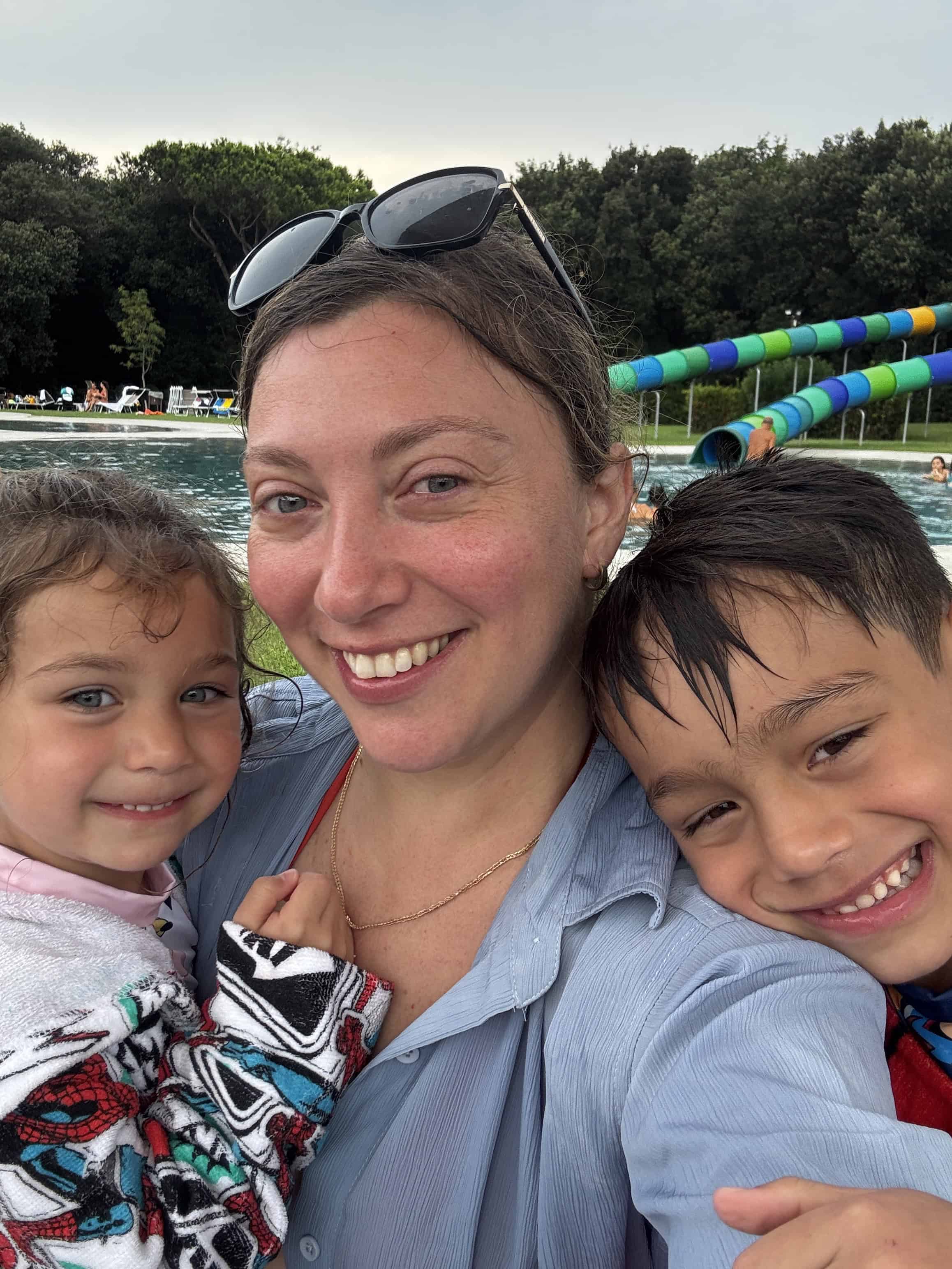 A woman smiles with two young children, all wet, at an outdoor pool with slides in the background&mdash;a perfect family travel moment for your bucket list.