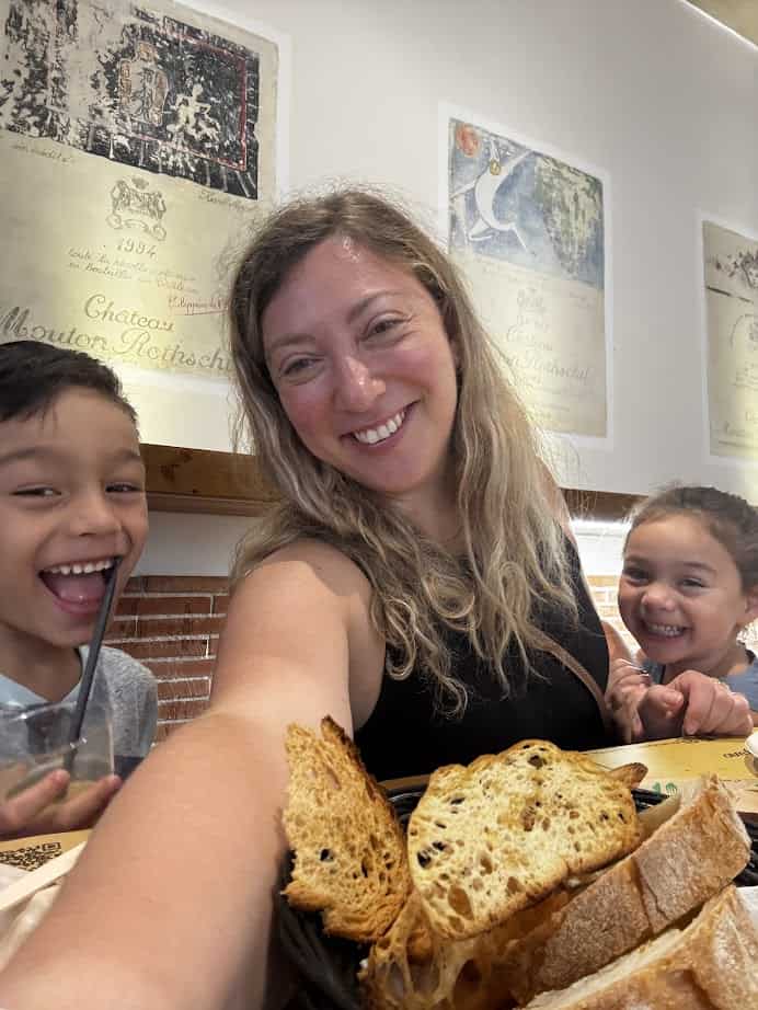 A woman and two kids smile at a table with toasted bread in a cozy restaurant with art on the wall, enjoying the chance to eat vegetarian in Italy.