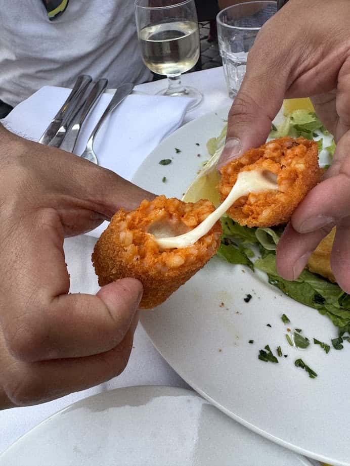 Two hands pull apart a fried croquette, revealing gooey melted cheese inside, over a plate of salad&mdash;a perfect way to eat vegetarian in Italy.