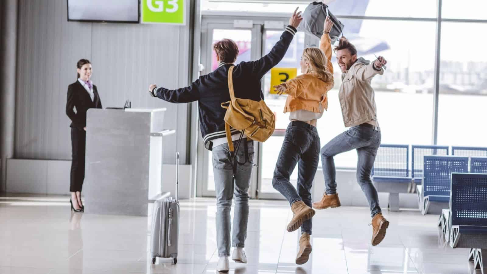 Three people with suitcases joyfully jumping in an airport terminal near gate G3, watched by a smiling staff member.