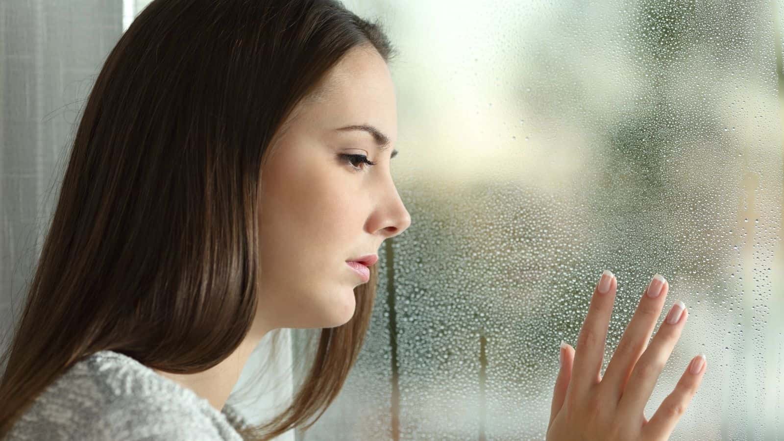A woman looks thoughtfully out a rain-covered window, touching the glass with her hand.
