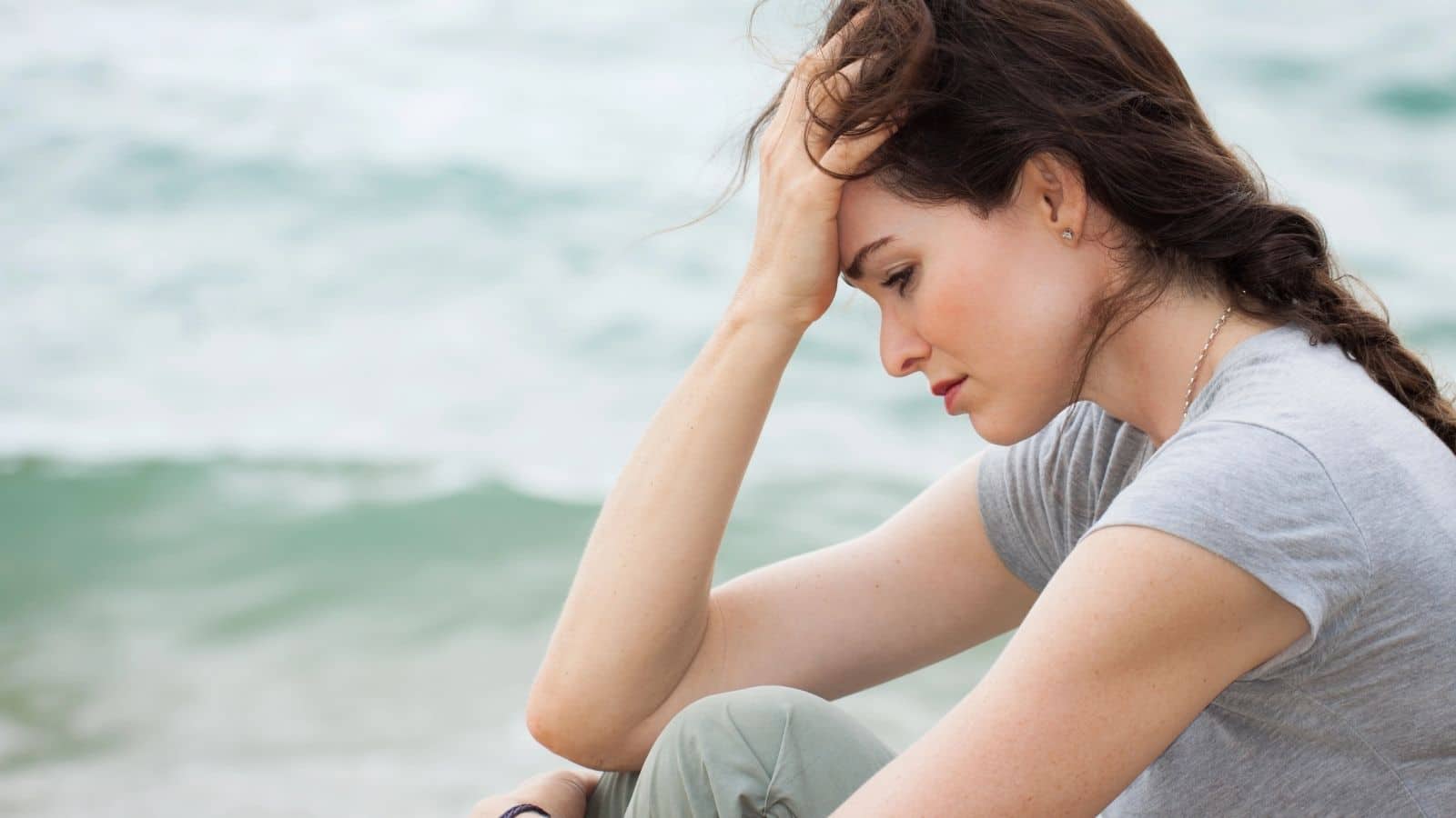A woman sits by the water, looking down with her hand on her forehead, appearing thoughtful or stressed.