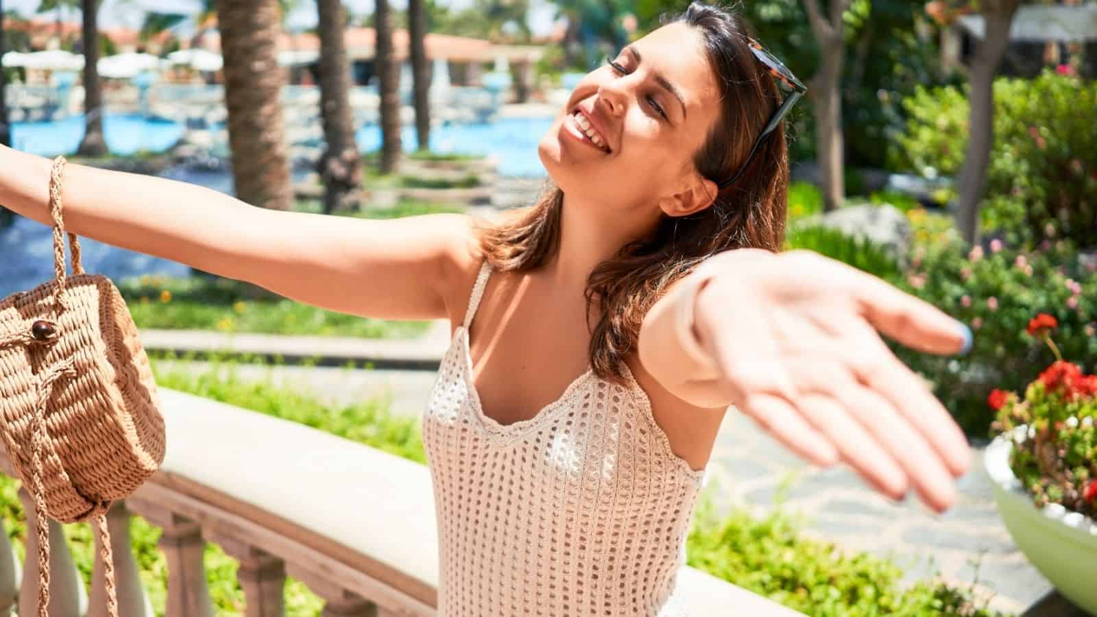 Woman in a white crochet dress smiling with open arms outdoors on a sunny day, holding a straw bag.