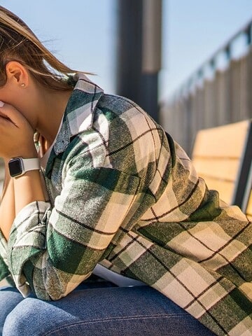 A woman sits on a bench at a train station, covering her face with her hands beside a purple suitcase.