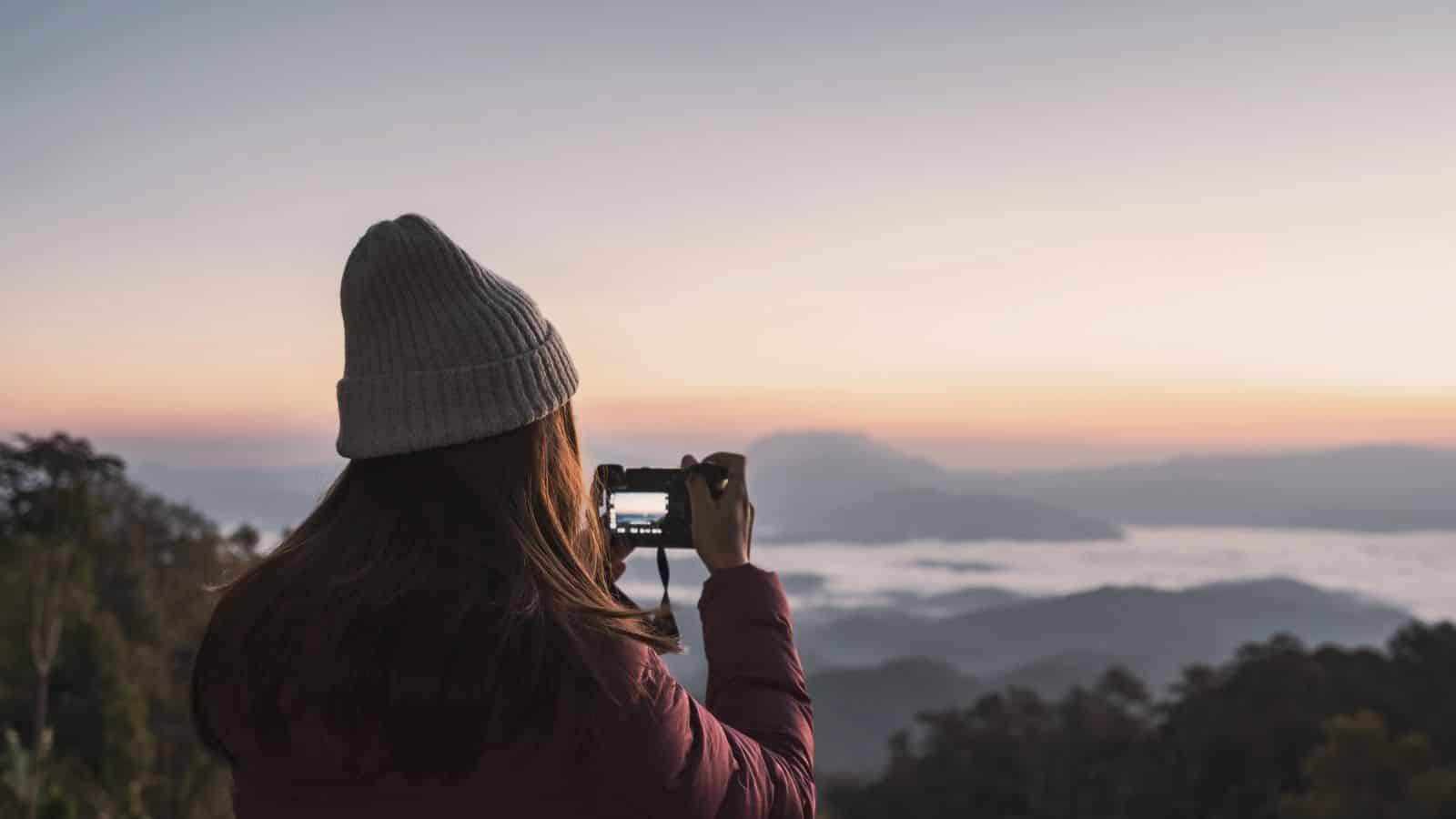 A person in a beanie takes a photo of a misty mountain sunrise with a camera.