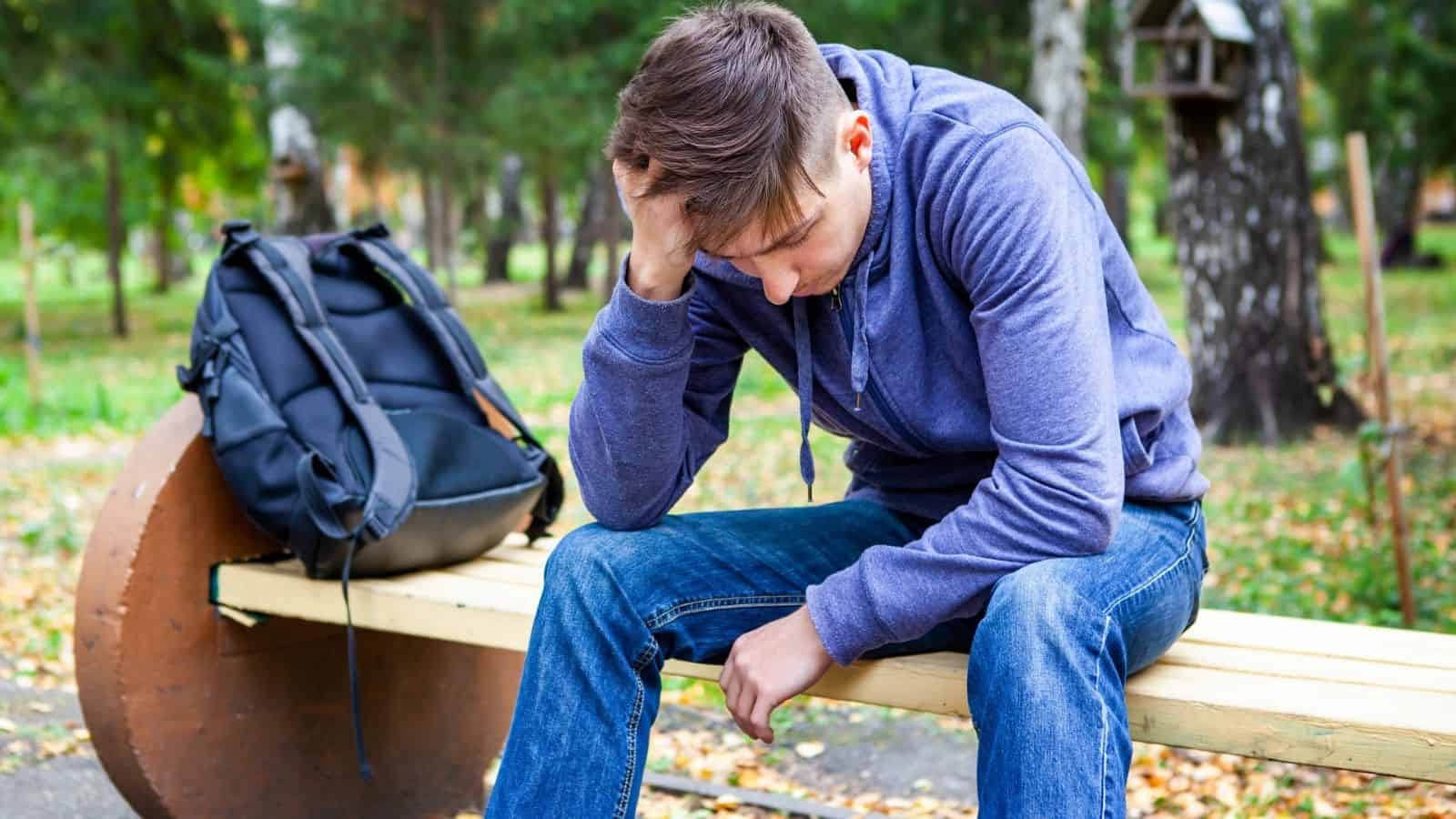 A young man sits on a bench outdoors, looking down with his hand on his head, appearing stressed.