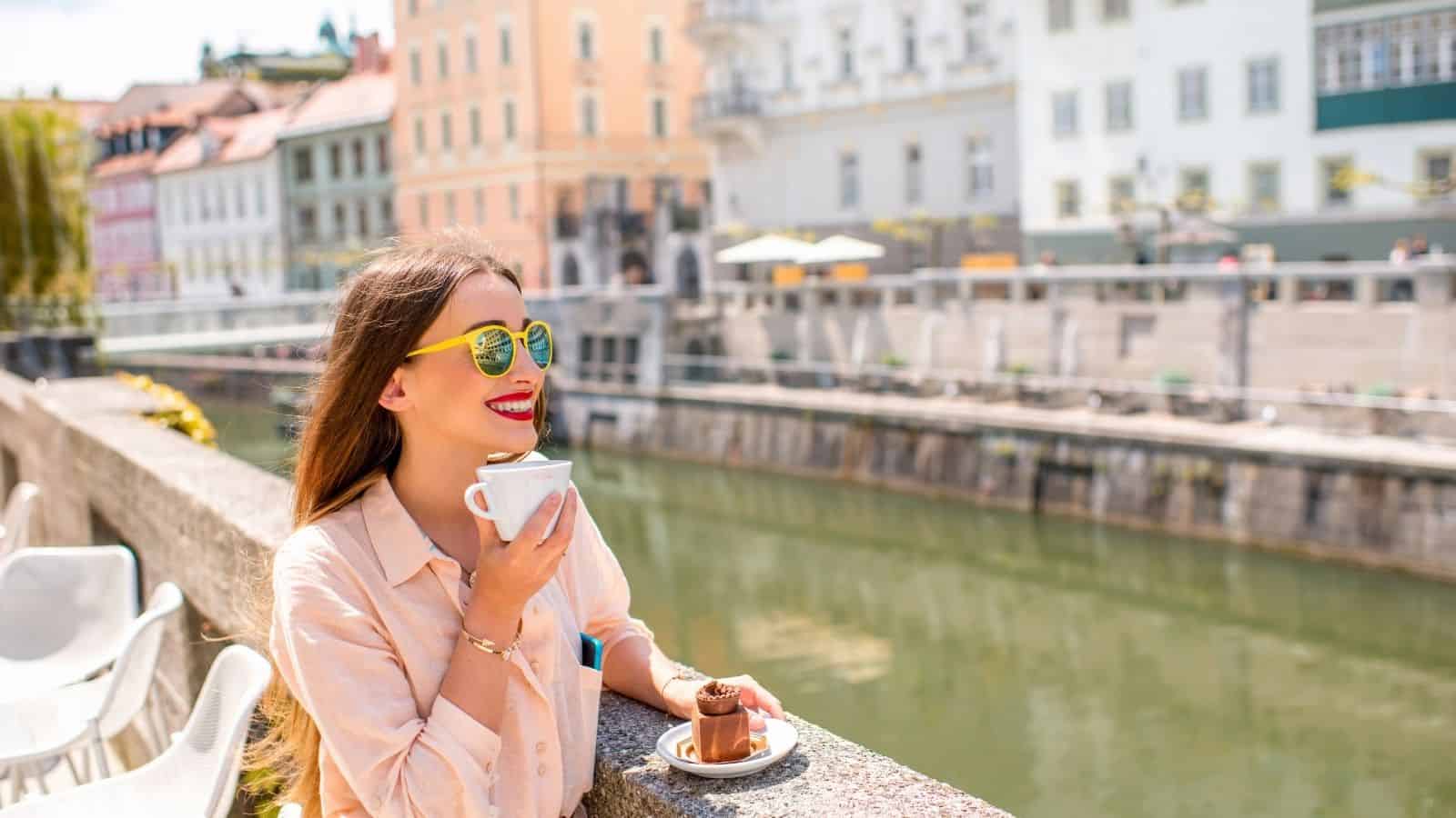 Woman in sunglasses sits by a riverside, smiling and drinking coffee with a dessert on the ledge.