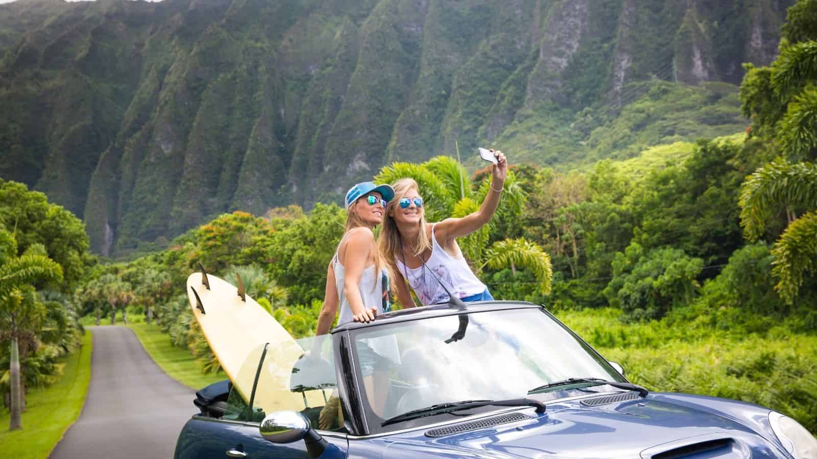 Two women in a convertible with a surfboard take a selfie on a lush, tropical road with mountains behind.