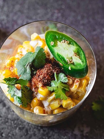 A glass of Mexican street corn, one of the Best Vegetarian Mexican Dishes, topped with chili powder, cilantro, and sliced jalapeño on a dark surface.