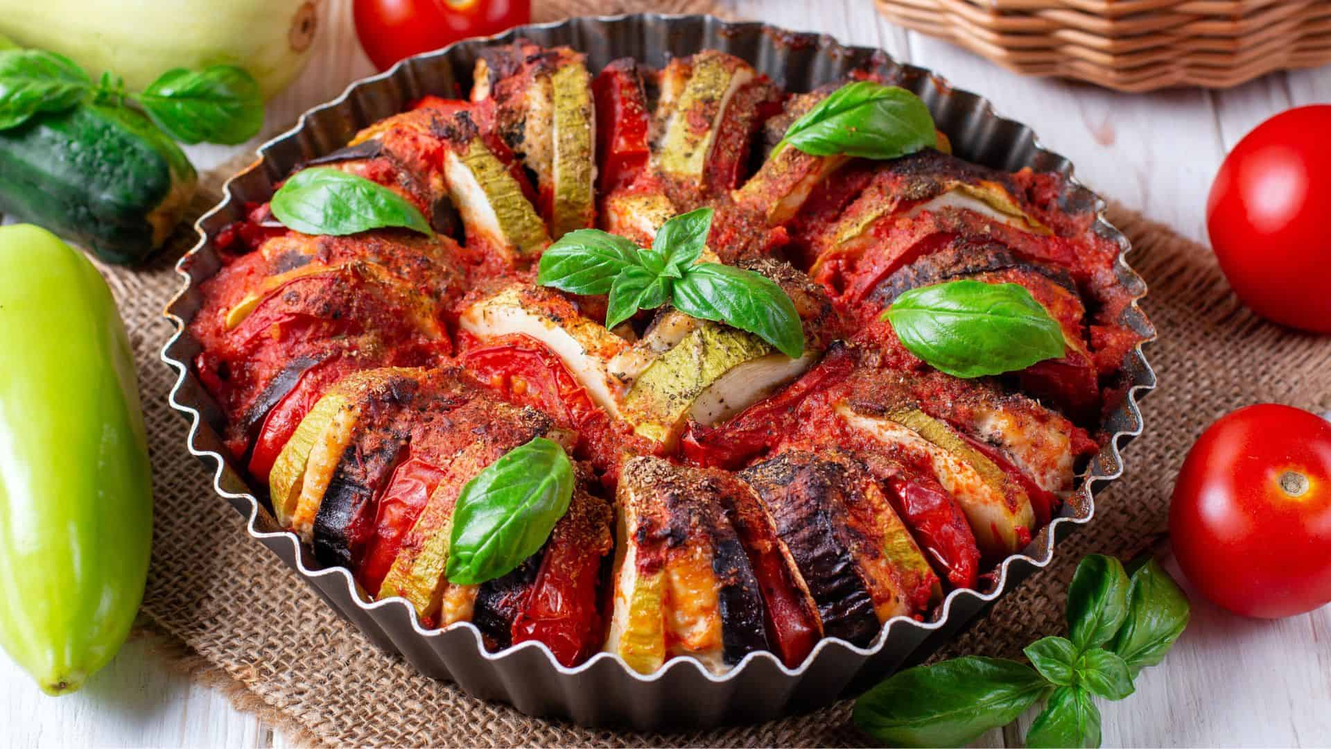A colorful baked ratatouille in a round pan, garnished with fresh basil and surrounded by fresh vegetables&mdash;a true classic among the Best Vegetarian Food in France.