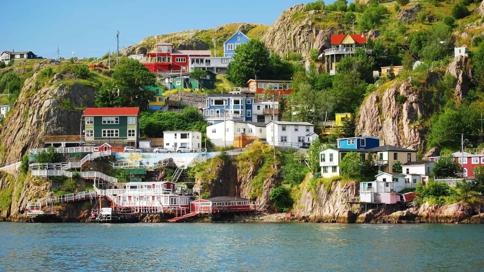 Colorful houses on a rocky hillside overlooking the water under a blue sky, surrounded by trees and greenery.