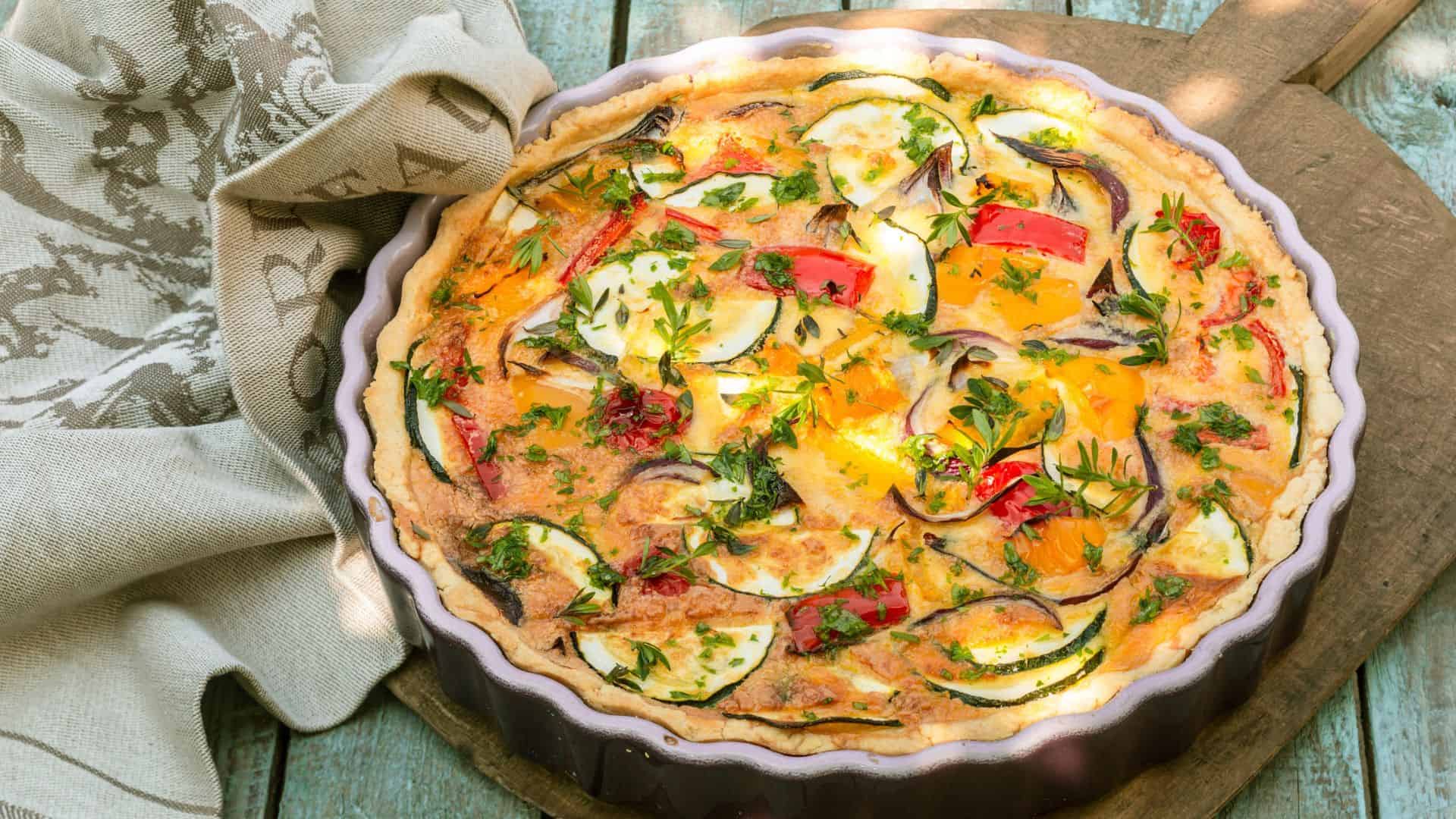Colorful vegetable quiche in a round dish on a wooden surface, next to a beige towel&mdash;a delightful example of the Best Vegetarian Food in France.