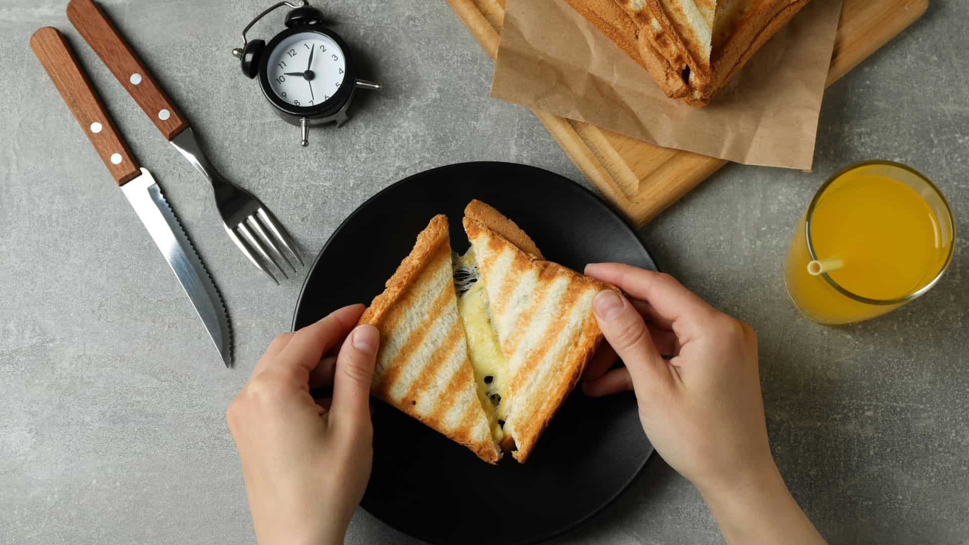 Hands holding a grilled sandwich over a black plate, with orange juice, cutlery, and a small clock nearby&mdash;capturing the essence of enjoying the Best Vegetarian Food in France.