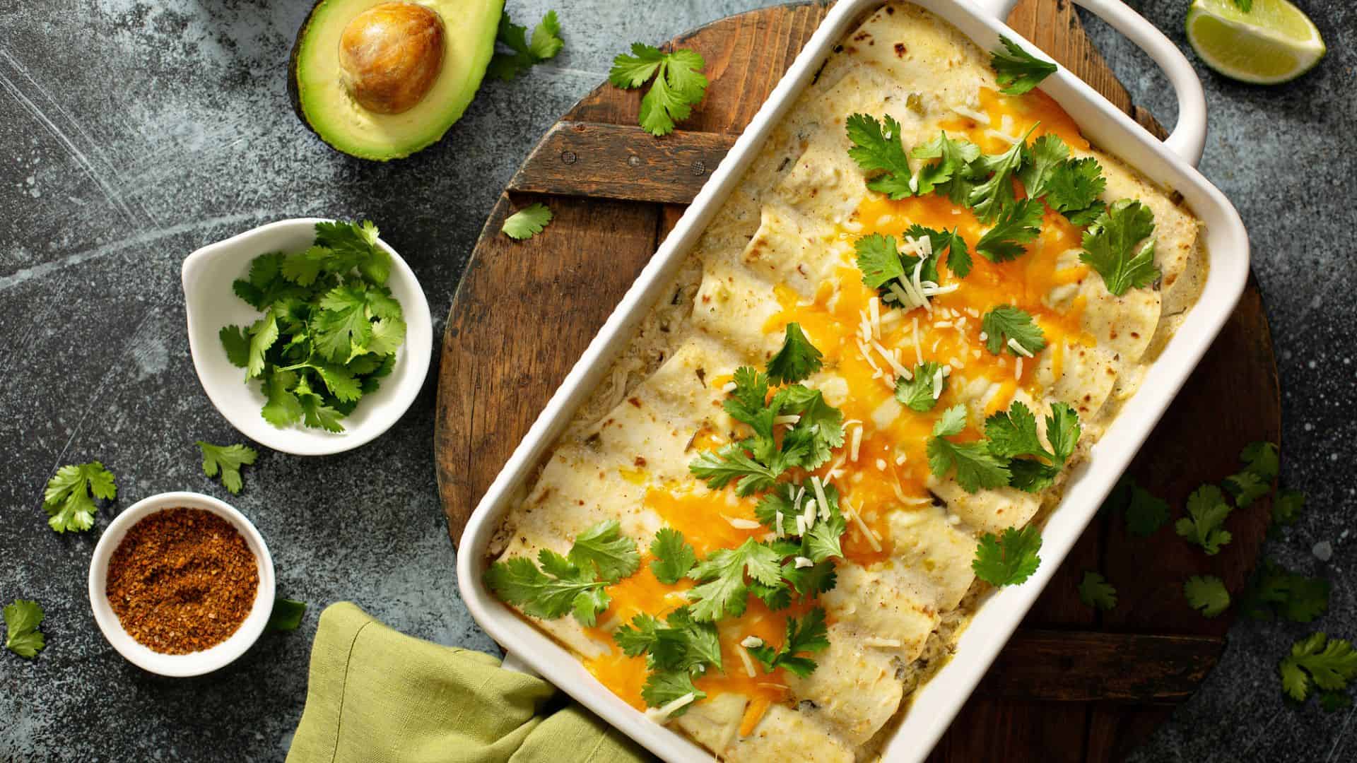 Baked enchiladas topped with melted cheese and cilantro in a white dish, surrounded by avocado and seasonings&mdash;one of the best Vegetarian Mexican Dishes for a delicious and vibrant meal.