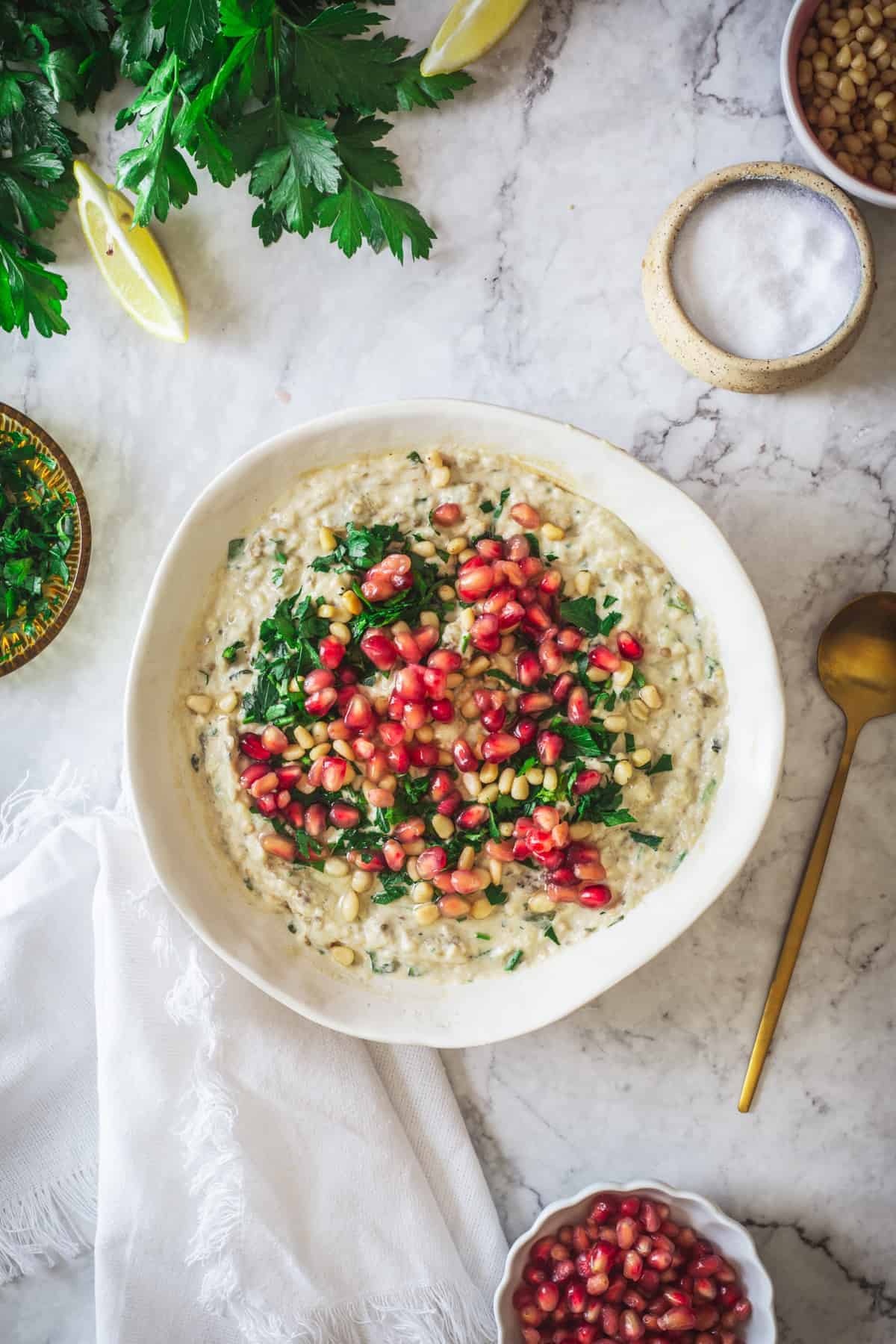 Baba ganoush topped with parsley, pine nuts, and pomegranate seeds in a white bowl on a marble surface&mdash;a delicious example of vegetarian food in Israel.