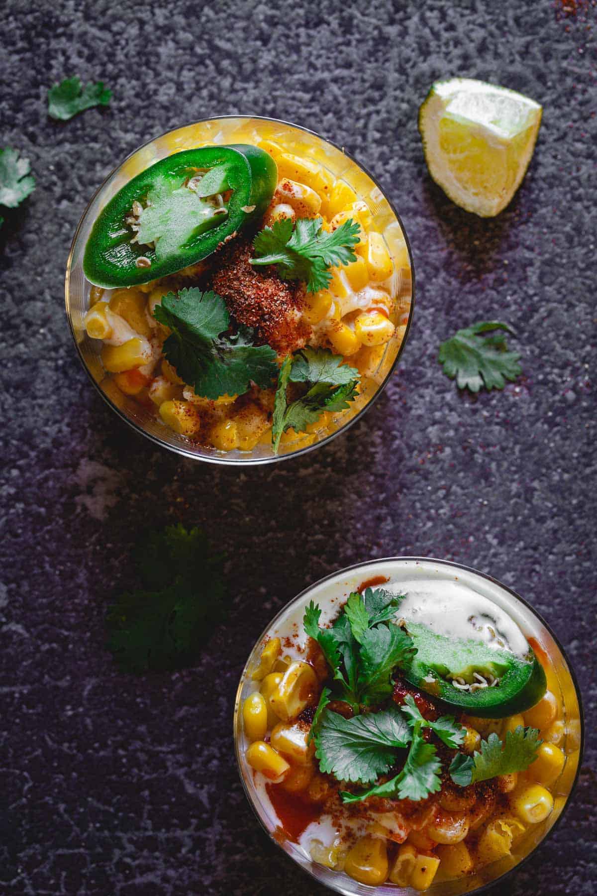 Two cups of Mexican street corn, one of the best vegetarian Mexican dishes, are topped with cilantro, jalape&ntilde;o slices, chili powder, and served with a lime wedge nearby.