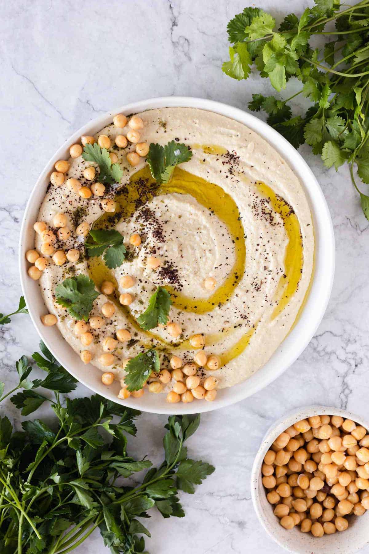 A bowl of hummus topped with olive oil, chickpeas, herbs, and spices&mdash;classic vegetarian food in Israel&mdash;surrounded by fresh parsley and cilantro.