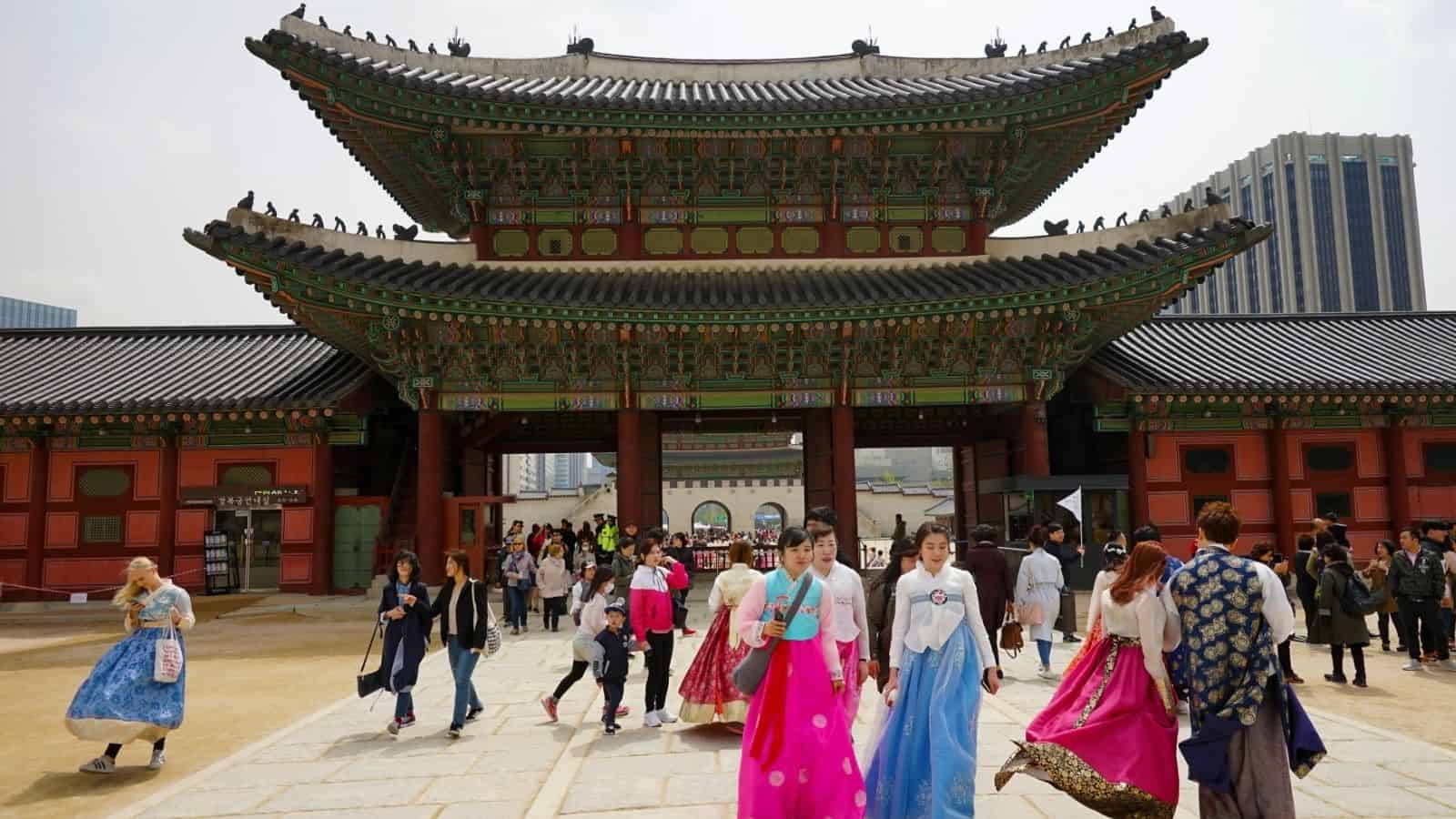 People in colorful traditional Korean hanbok walk in front of a large ornate palace gate with others nearby.