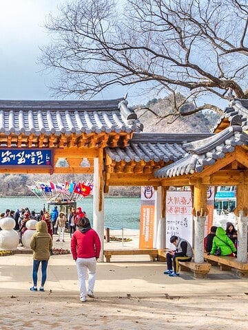 People walk near a traditional Korean pavilion by a river, with mountains and trees in the background.