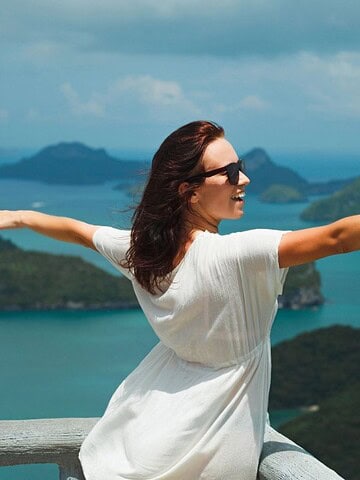 Woman in a white dress poses with arms outstretched, overlooking tropical islands and blue sea—embracing hot weather travel with ease.