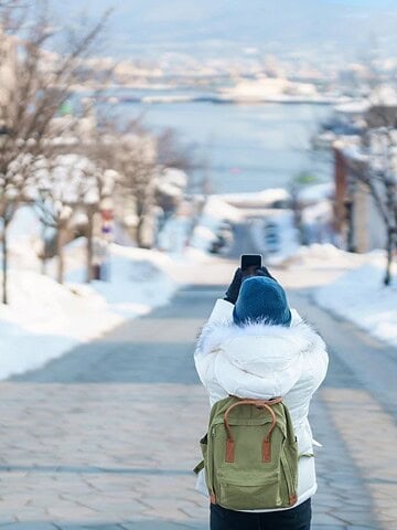 Person in winter clothes taking a photo on a snowy street with water and mountains in the background.