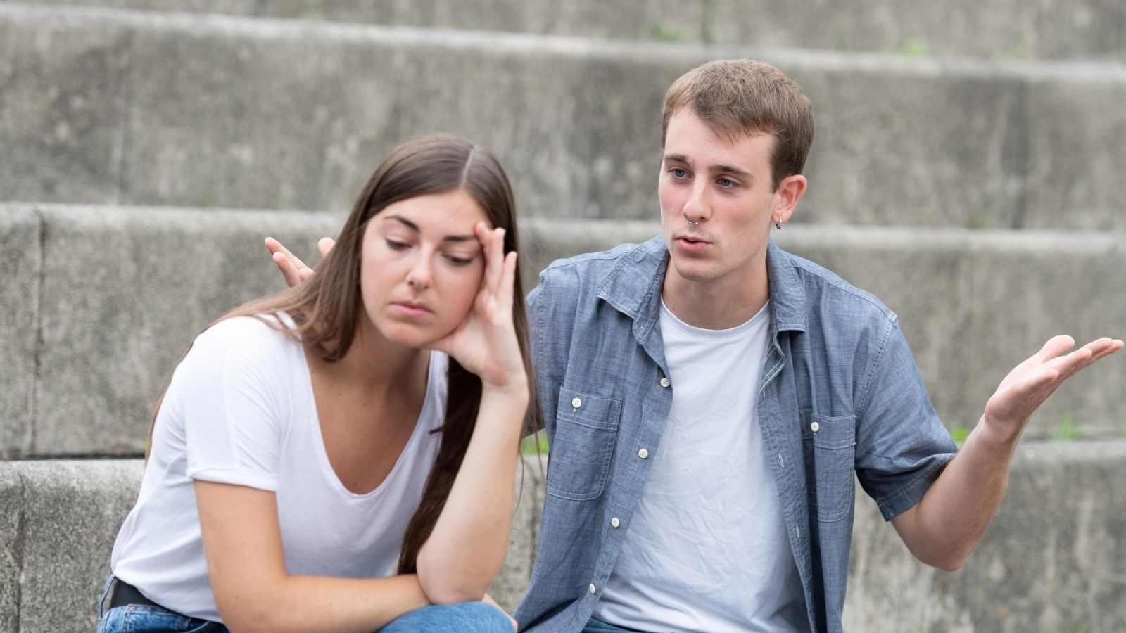 A young woman looks upset while a young man gestures as if explaining something on outdoor steps.