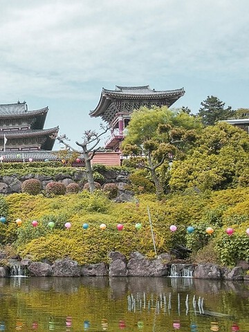 People walk near a pond toward traditional temples decorated with colorful lanterns and surrounded by greenery.