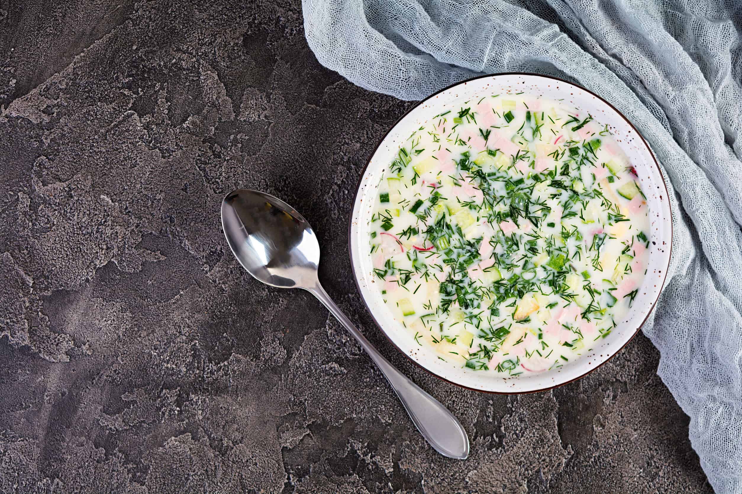 A bowl of cold soup with herbs, diced vegetables, and ham next to a spoon on a textured surface&mdash;this classic dish is a refreshing counterpart to popular vegetarian dishes in Russia.