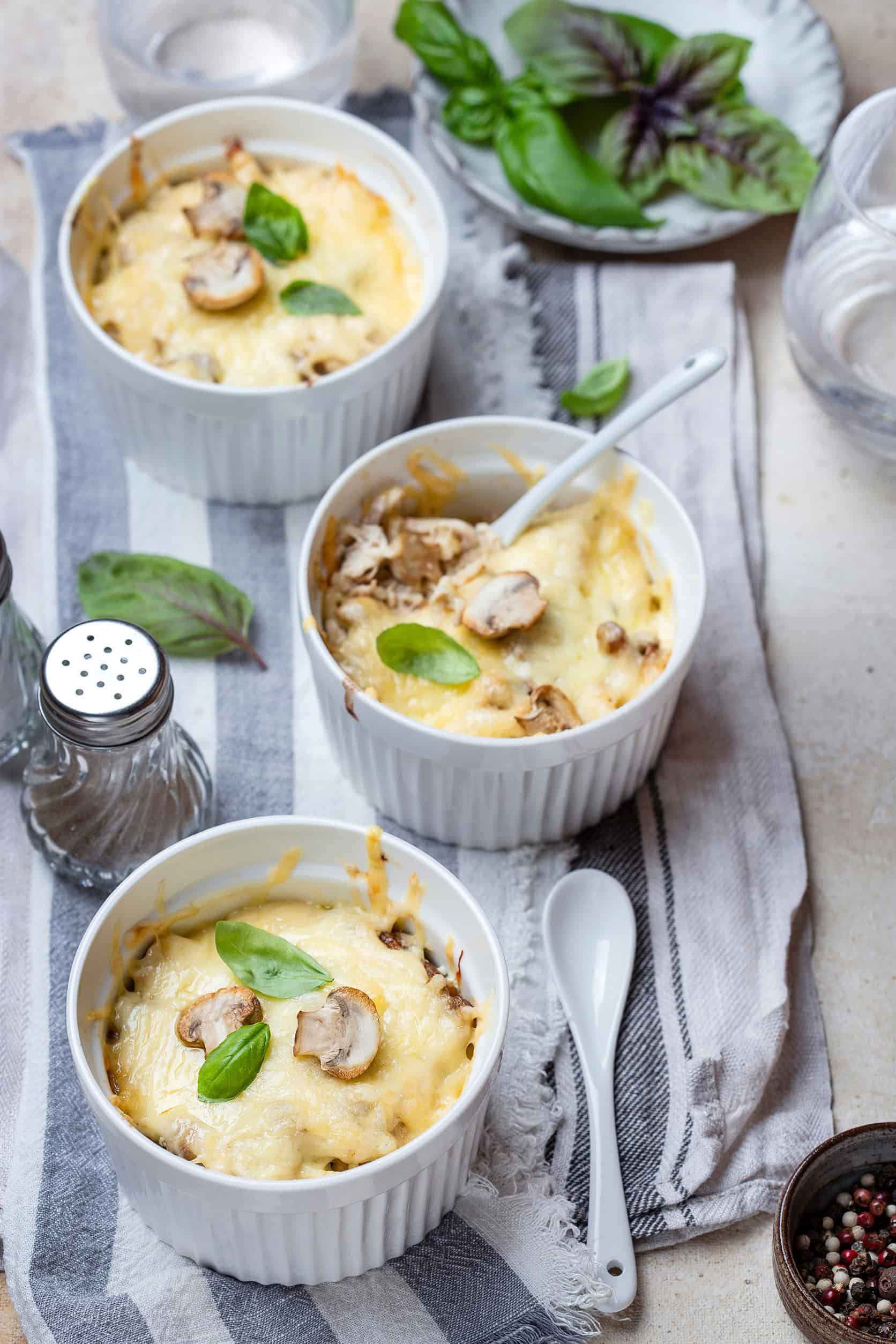 Three ramekins of baked cheesy casserole with mushrooms and basil leaves on a striped cloth, showcasing one of the delicious vegetarian dishes in Russia, served with spoons and water.