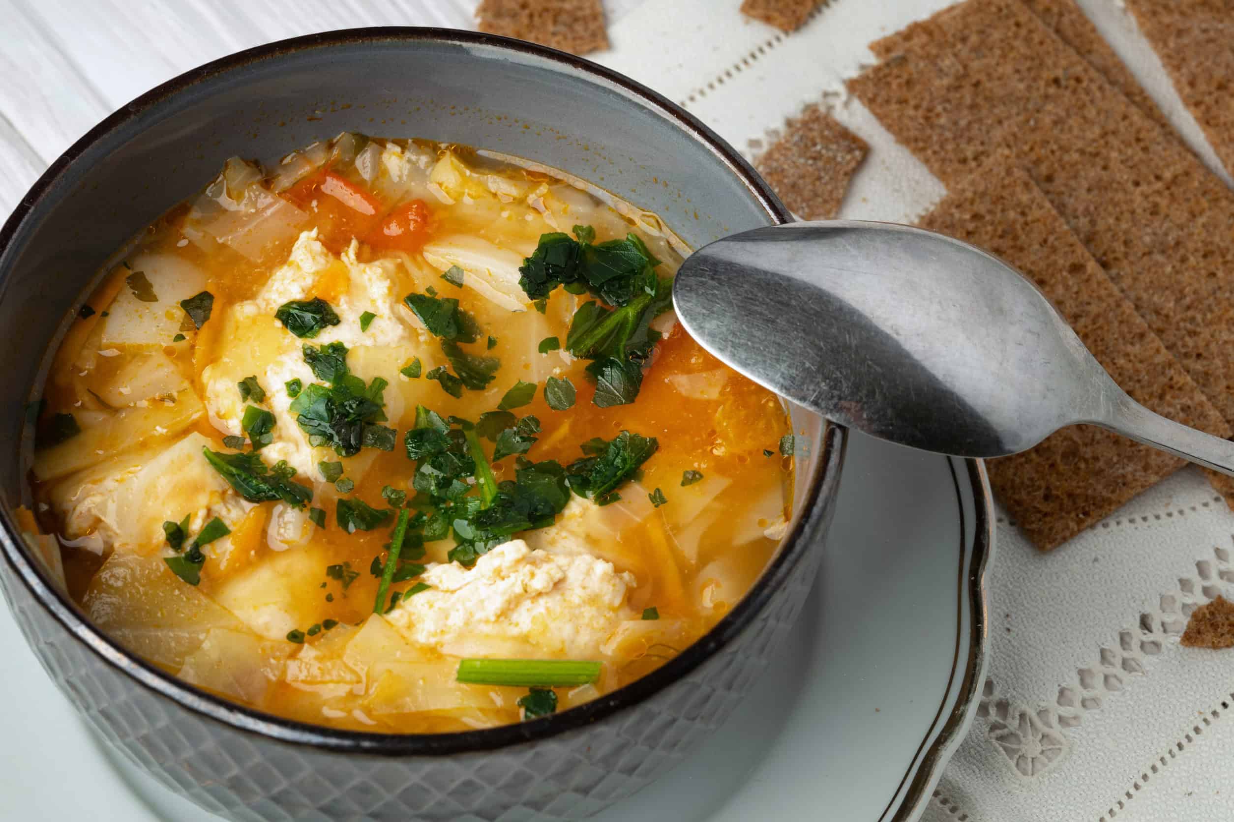 A bowl of vegetable soup with herbs and cheese, a classic among vegetarian dishes in Russia, sits next to a spoon and pieces of crispbread on a white tablecloth.