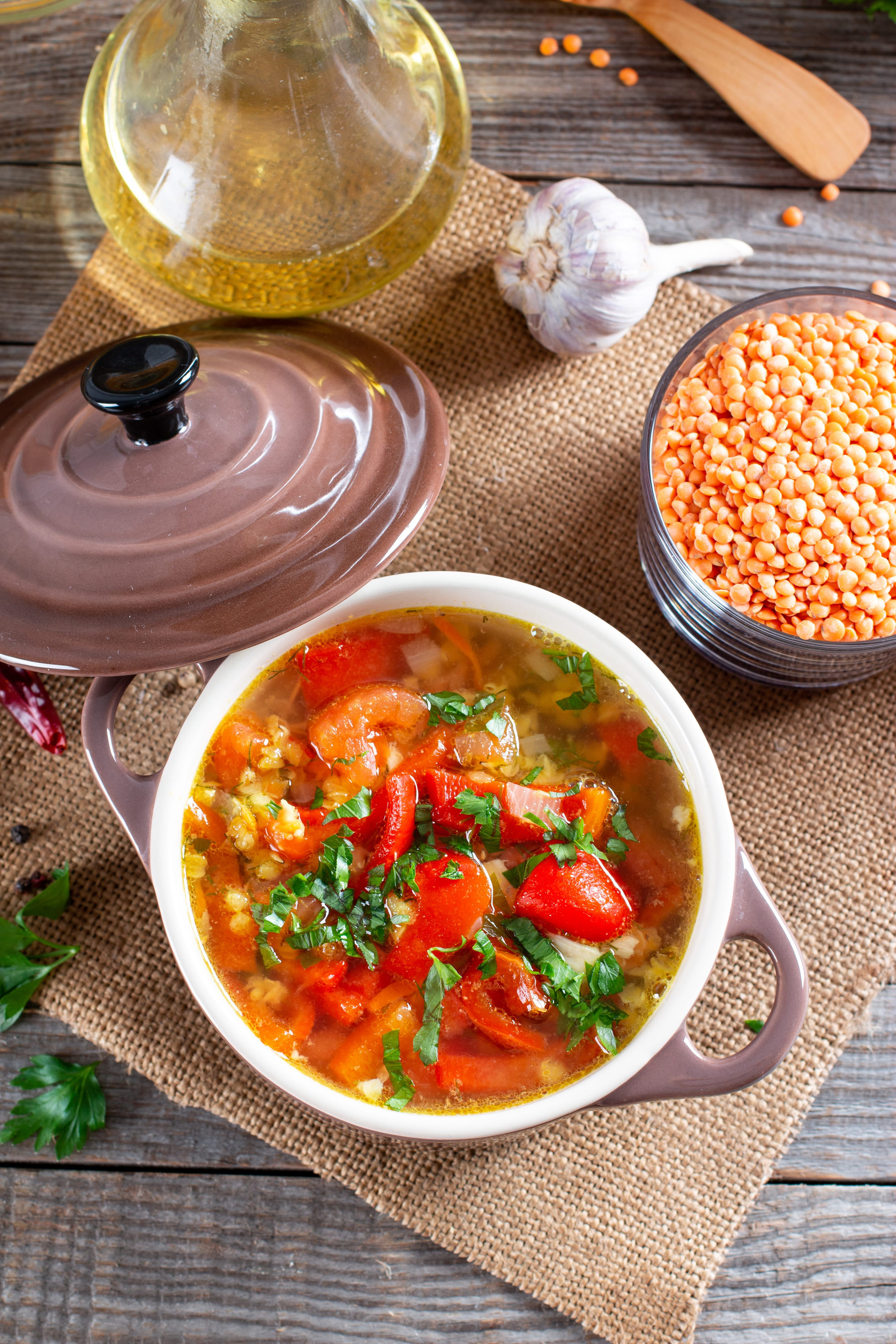 A bowl of lentil soup with tomatoes and herbs, a classic among vegetarian dishes in Ukraine, sits next to dry lentils, garlic, and oil on a rustic table.