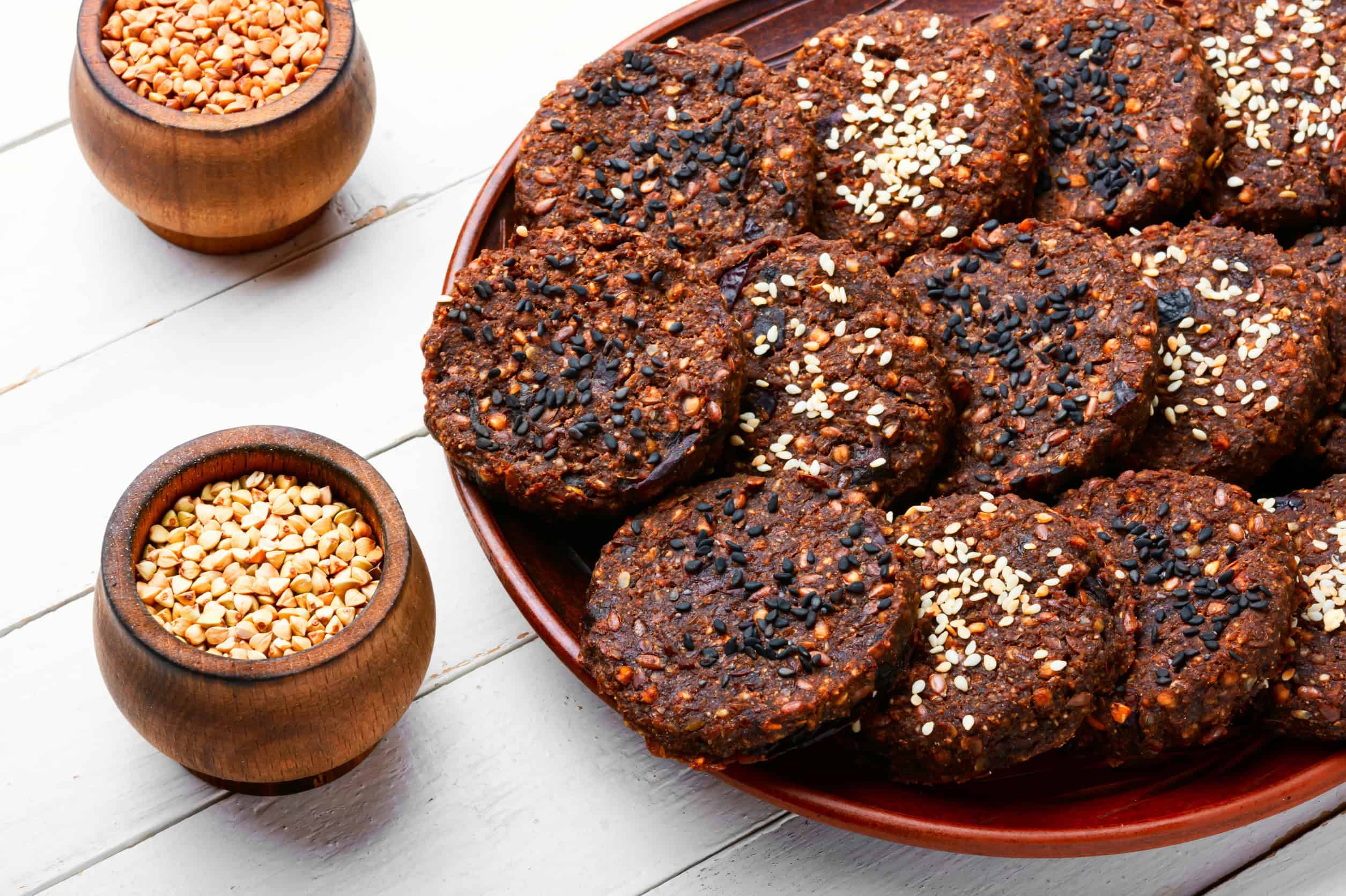 A plate of dark cookies topped with sesame seeds, next to bowls filled with buckwheat and grains on a white table, showcases delicious Ukrainian vegetarian dishes.