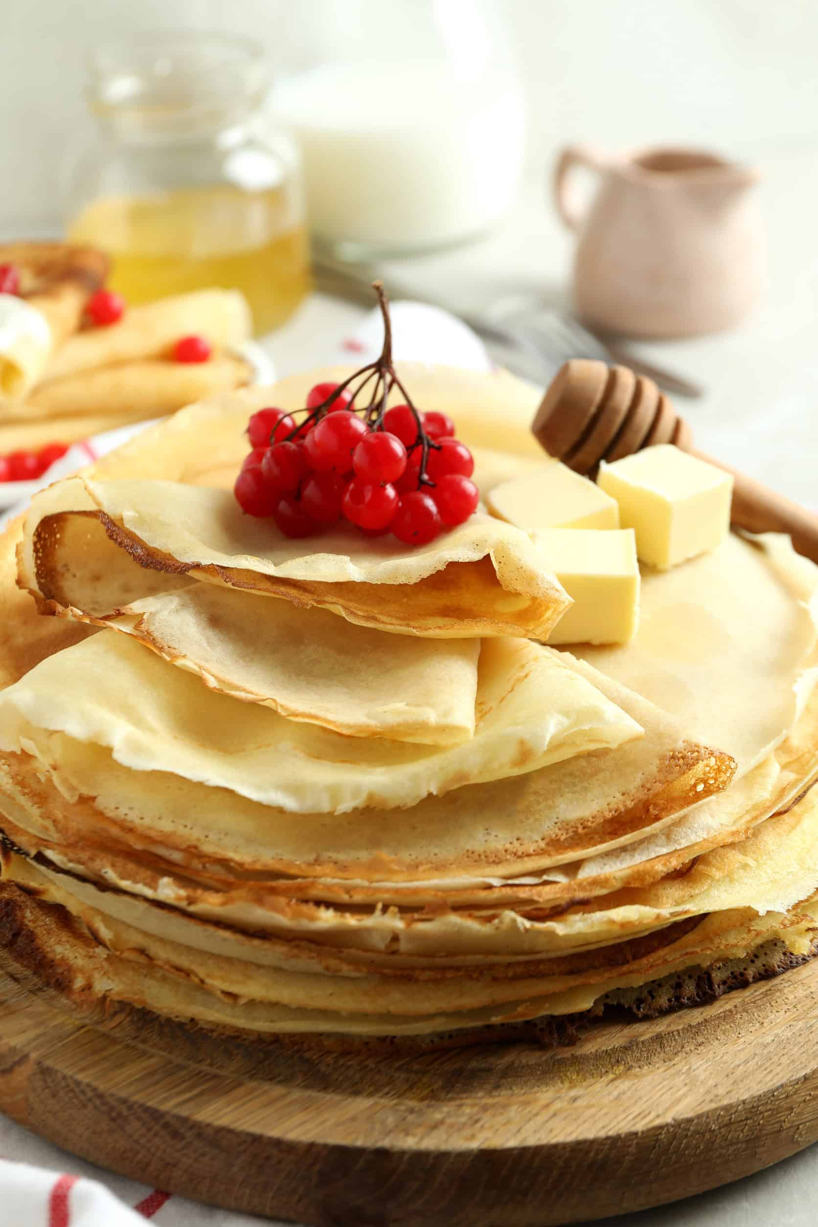 A stack of crepes with butter, red berries, and a honey dipper on a wooden board&mdash;a classic example of vegetarian dishes in Russia.