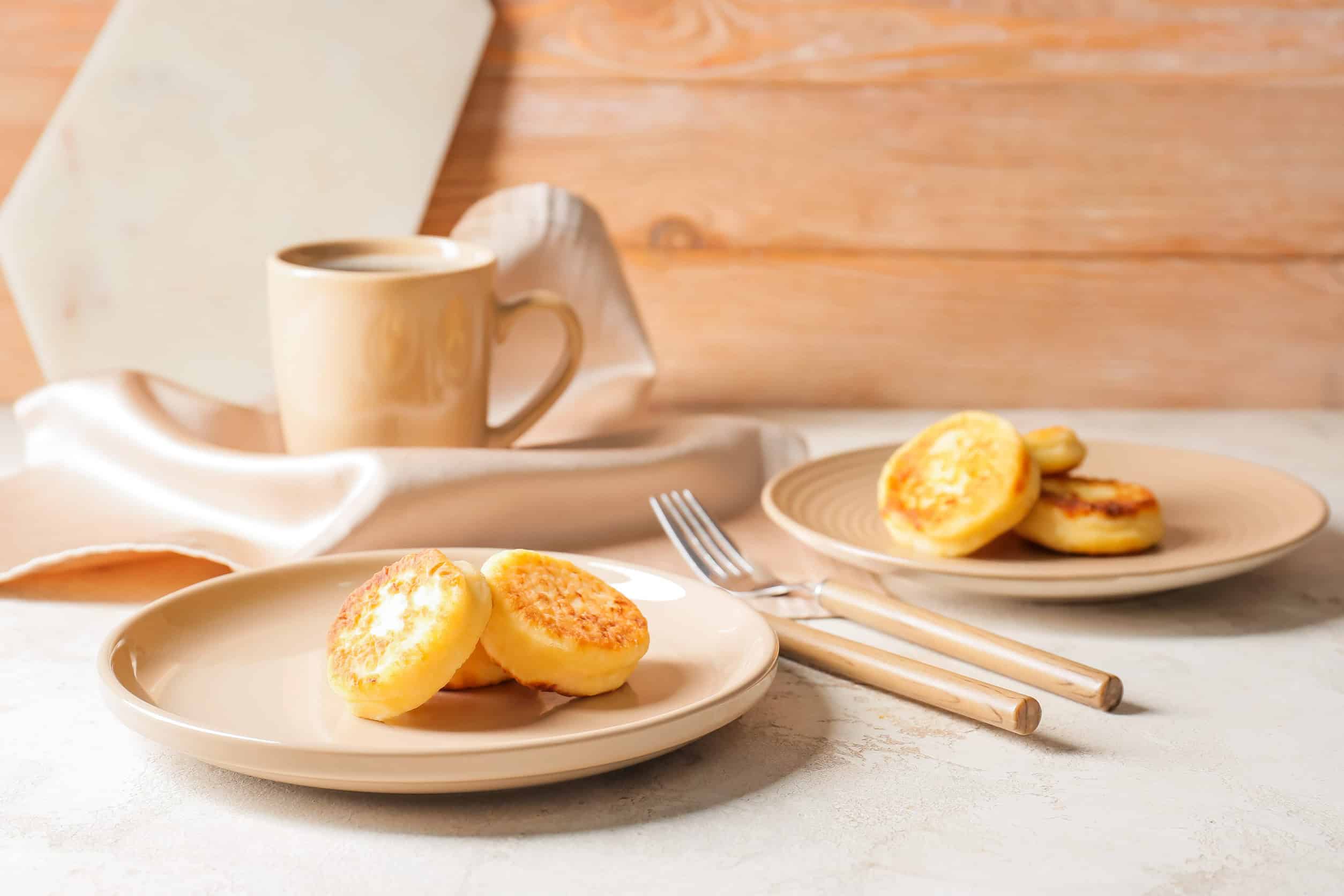 Two beige plates with pancakes, a popular choice among vegetarian dishes in Ukraine, sit beside a cup of coffee and utensils on a light table with a wooden background.