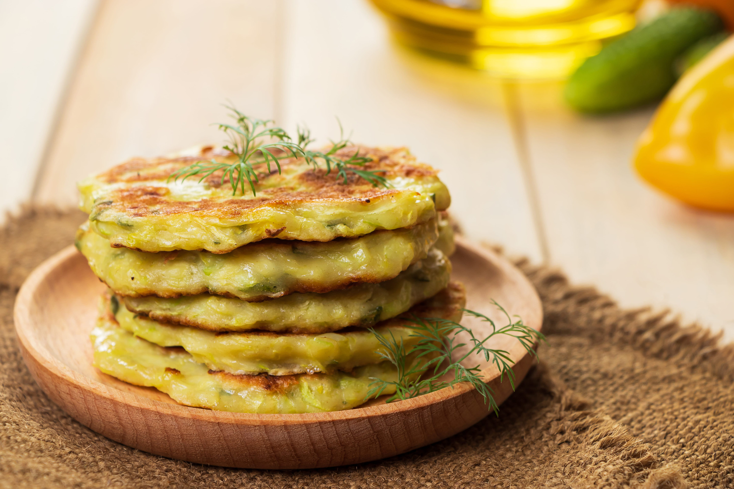 Four zucchini fritters stacked on a wooden plate, garnished with fresh dill on a rustic table&mdash;a delightful nod to Ukrainian cuisine and beloved vegetarian dishes.