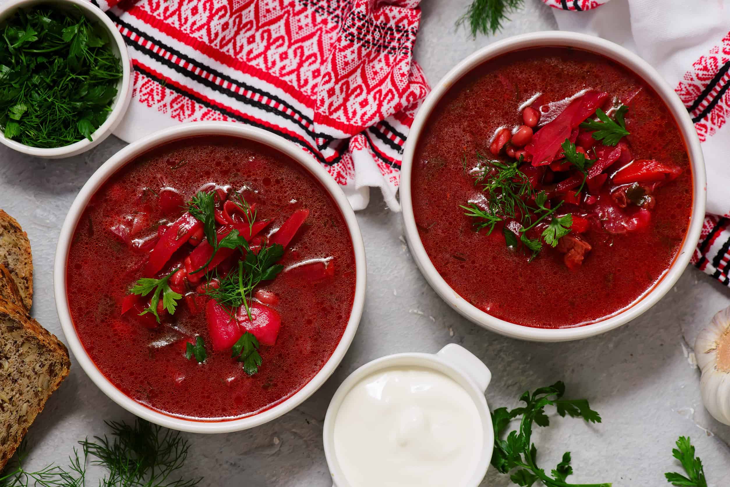 Two bowls of red borscht soup with herbs and sour cream, served alongside bread and a traditional embroidered cloth&mdash;a classic from Ukraine often praised among the top 13 vegetarian dishes.