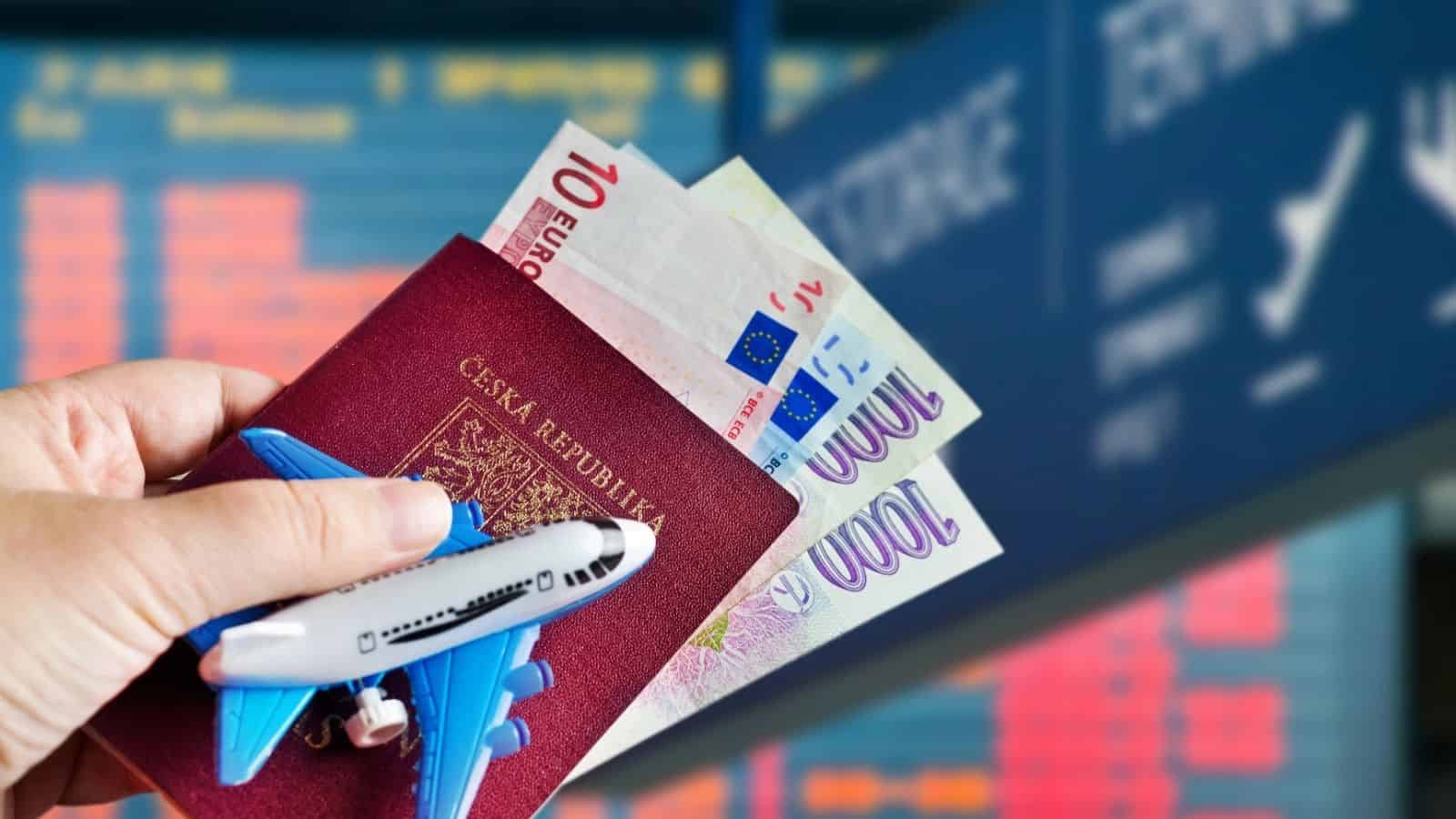 Hand holding a Czech passport, euro bills, and a toy airplane in front of a flight information board.