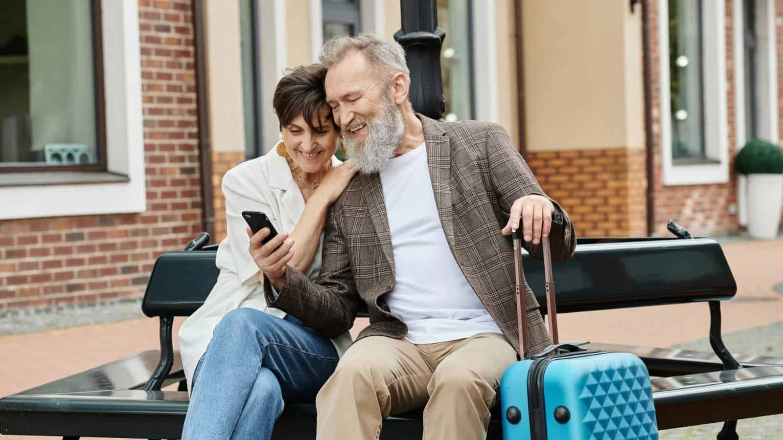 Smiling older couple sits on a bench with a blue suitcase, looking at a smartphone together.