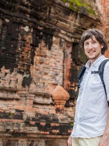 Man with backpack smiling in front of ancient brick ruins with green plants nearby.