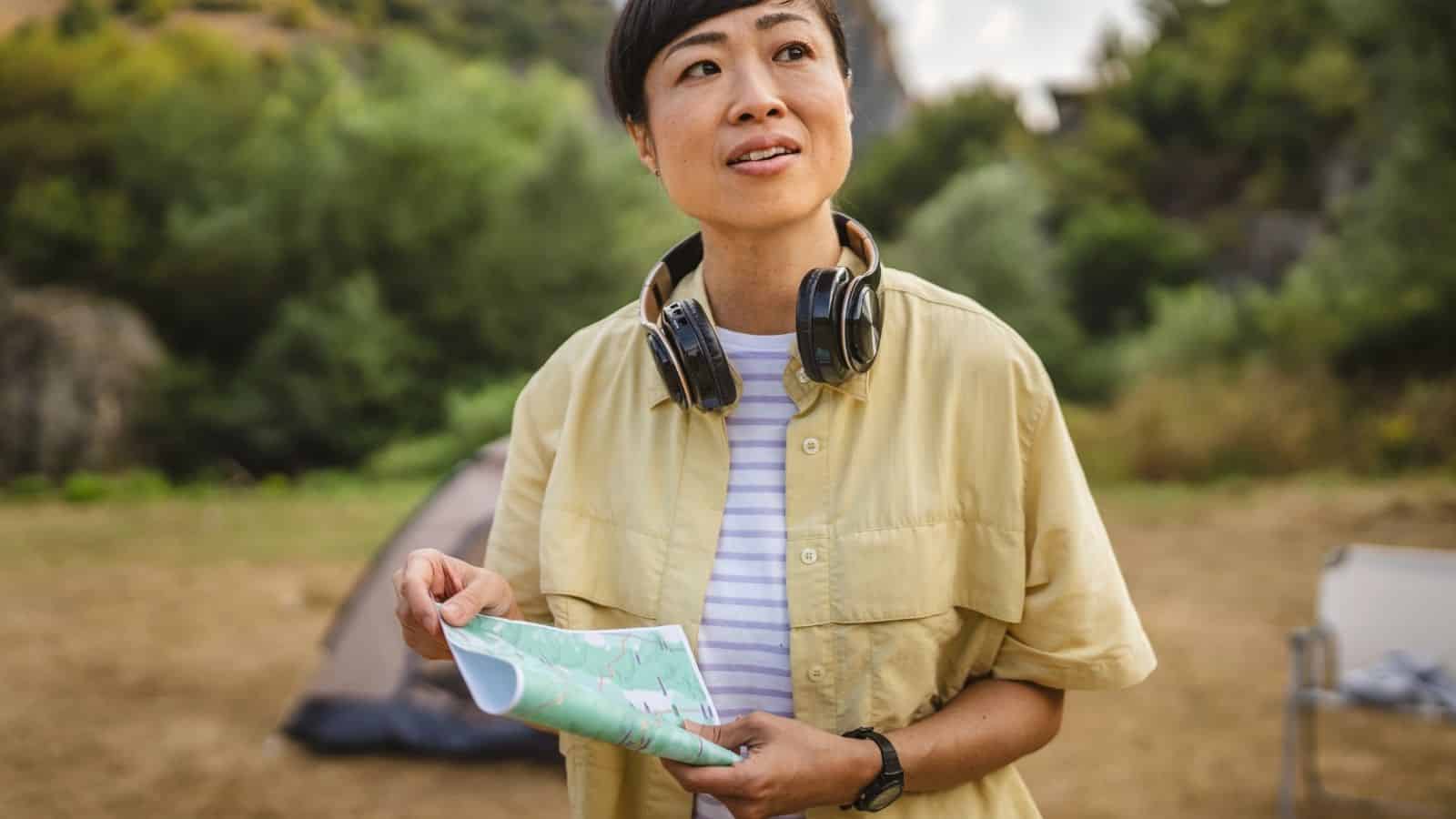 A person outdoors holding a map, wearing headphones around their neck, with a tent and trees in the background.
