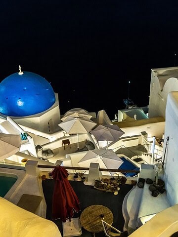 Night view of white buildings and blue domes in Santorini, Greece, with lights and umbrellas visible.