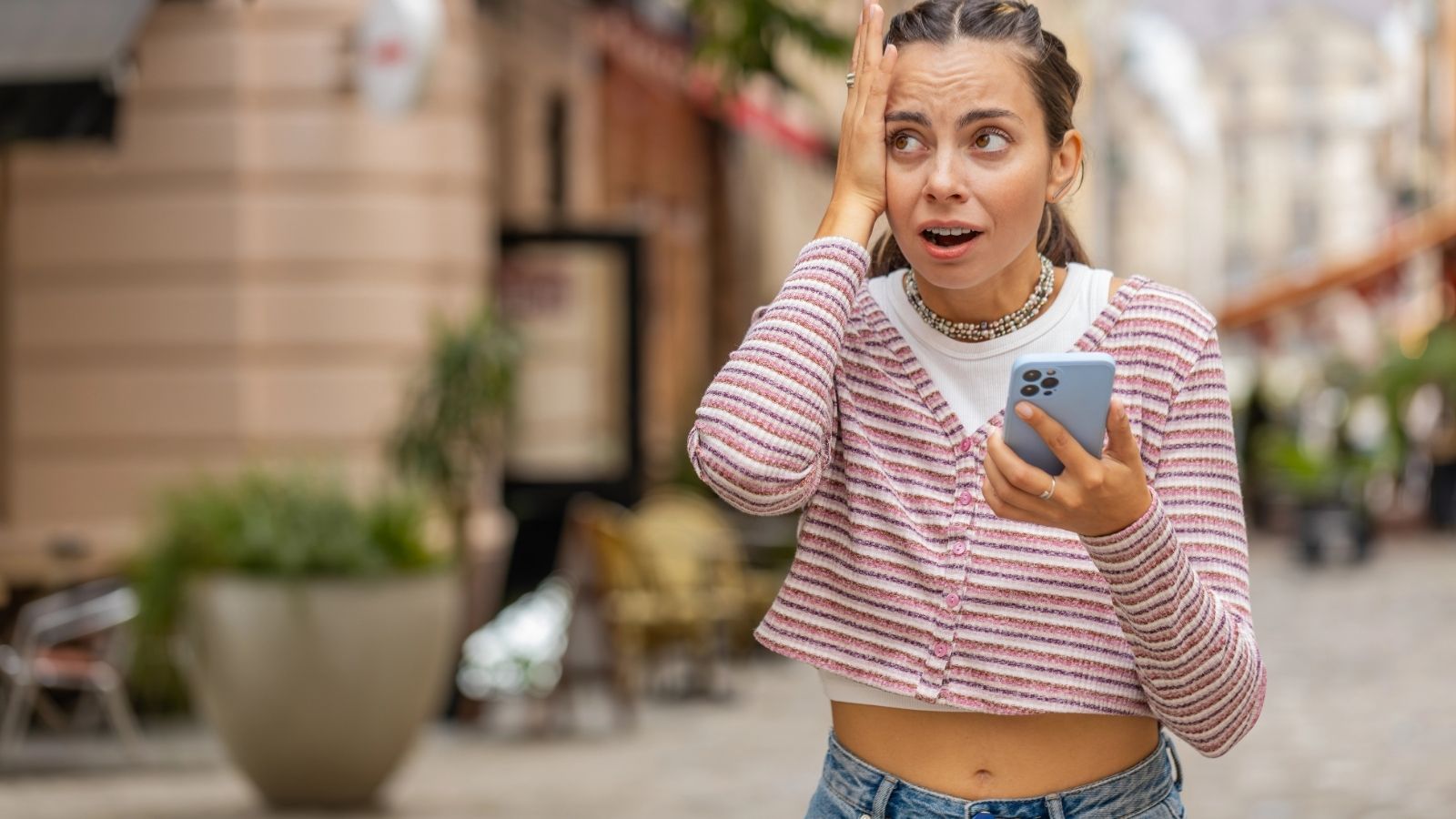 A shocked woman holds her phone and hand to her forehead outside.