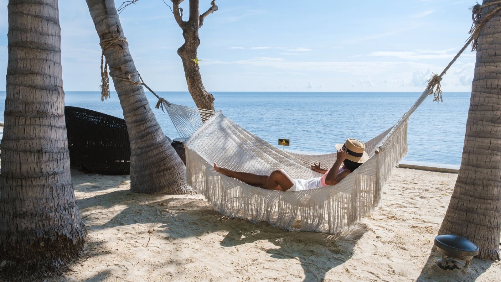 Person relaxing in a hammock tied between palm trees on a sandy beach by the ocean, wearing a hat—perfect for hot weather and ideal for anyone seeking Asia travel hacks.