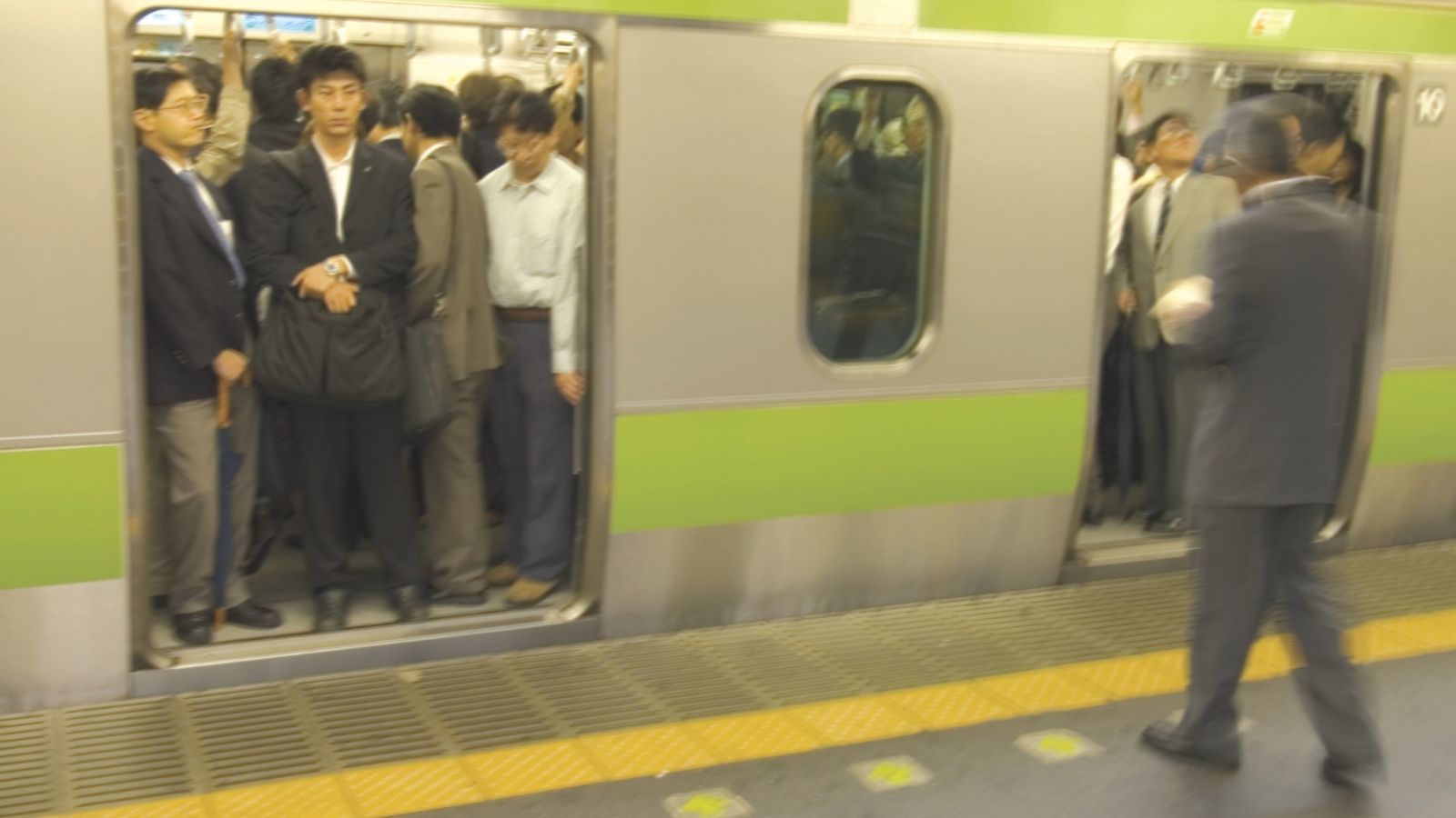Commuters in suits crowded inside a train with doors open at a station; one person stands on the platform.