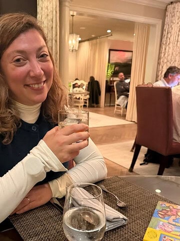 Woman smiling and holding a drink at a restaurant table with a glass of water and menus nearby, enjoying a toast after touring the best wineries in Finger Lakes.
