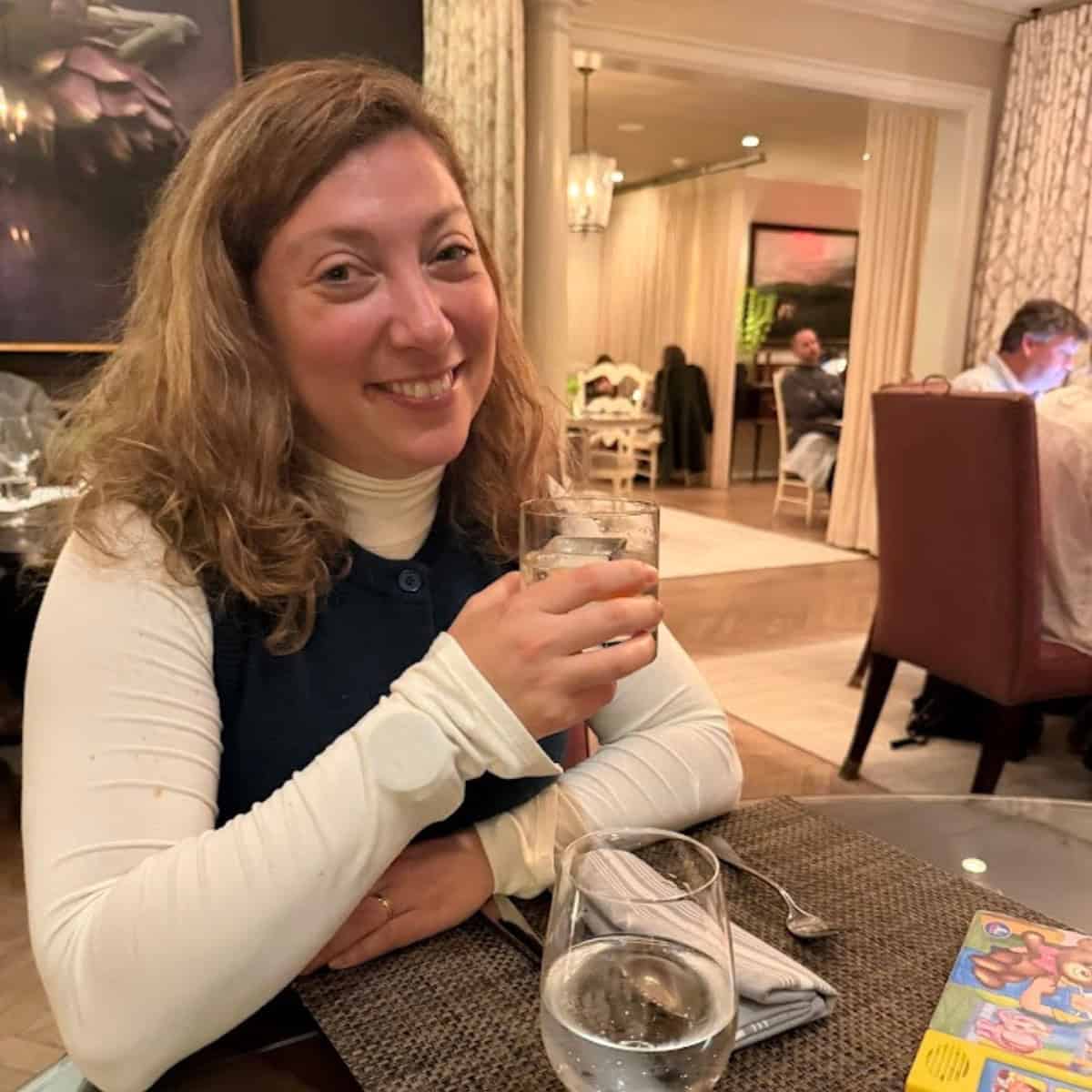 Woman smiling and holding a drink at a restaurant table with a glass of water and menus nearby, enjoying a toast after touring the best wineries in Finger Lakes.