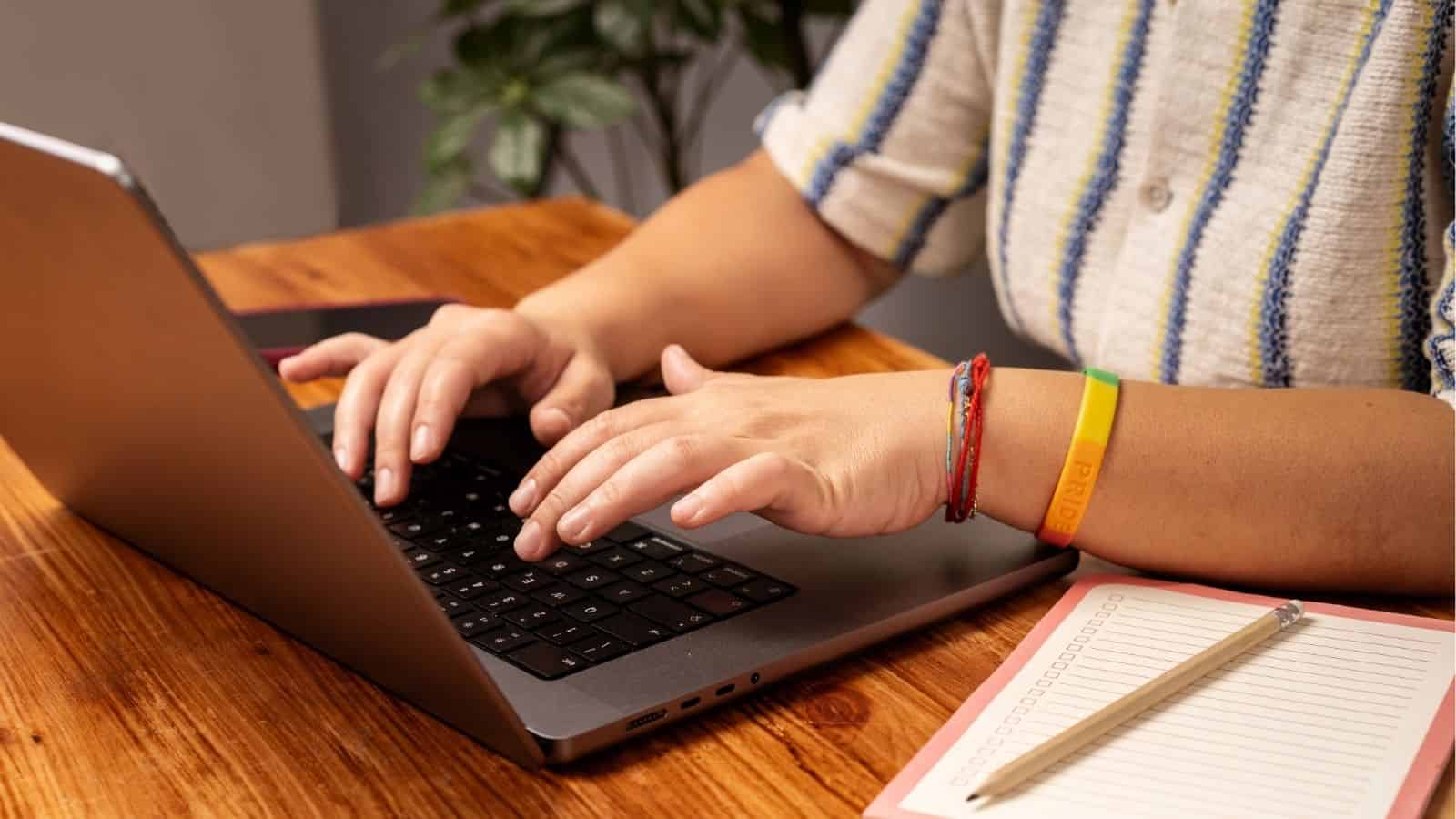 Person typing on a laptop at a wooden desk with a notepad, pencil, and potted plant in the background.