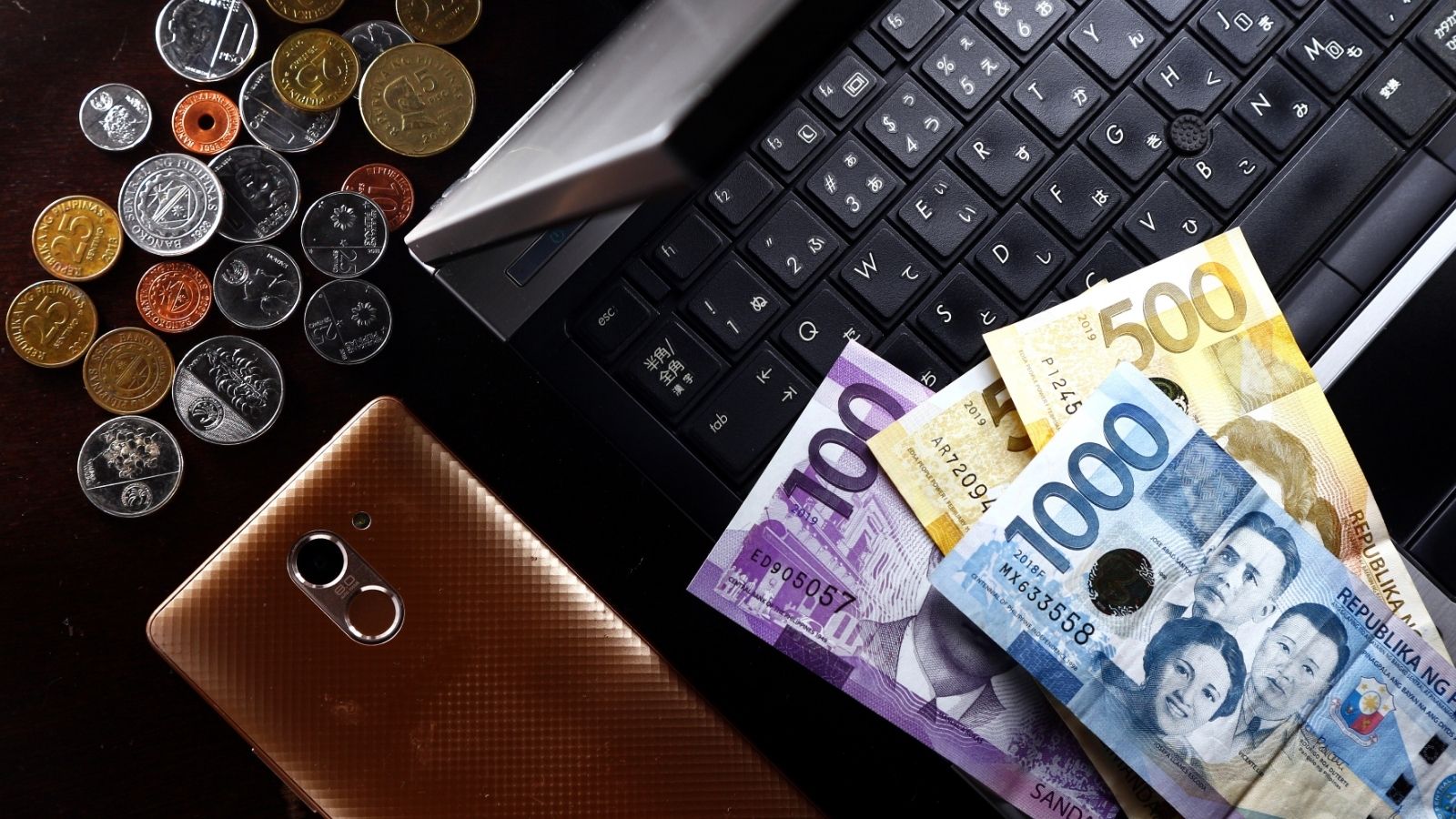 Philippine bills and coins, a brown smartphone, and a laptop keyboard on a dark table—essentials for Asia travel hacks and useful travel tips, especially when planning for hot weather adventures.